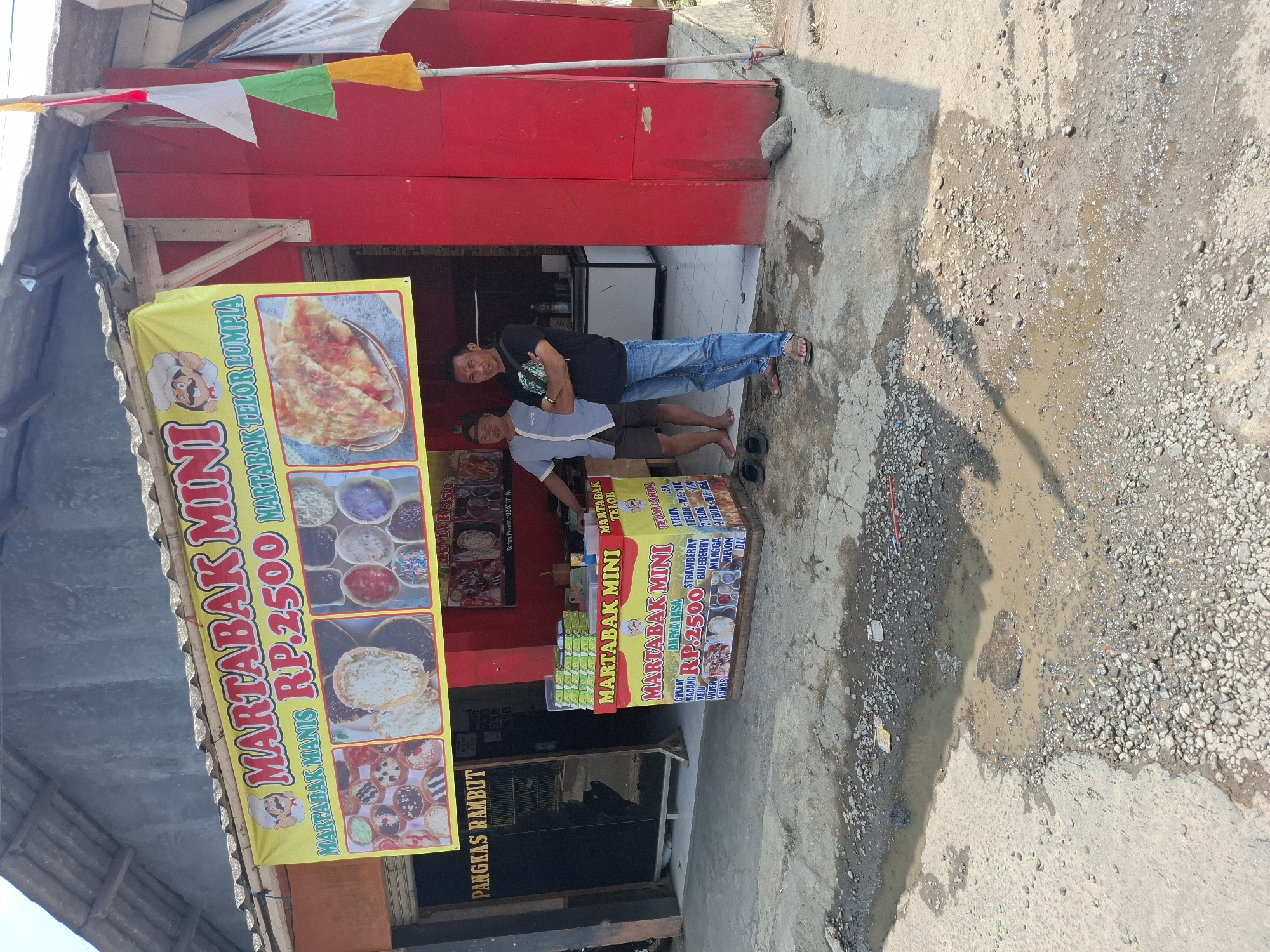 A Martabak Mini stall is set up in front of a building in Karangtengah, Indonesia. The stall is covered by a yellow banner advertising "MARTABAK MINI" with pictures of martabak and the price "RP.2500". A red and white Indonesian flag is attached to a pole on the right side of the stall. Two men are standing in front of the stall, one wearing a blue and white shirt and grey shorts, and the other wearing a black t-shirt and blue jeans. The ground in front of the stall is paved with concrete and gravel, with several puddles of water. The building behind the stall appears to be a shop with a red facade and a grey roof.