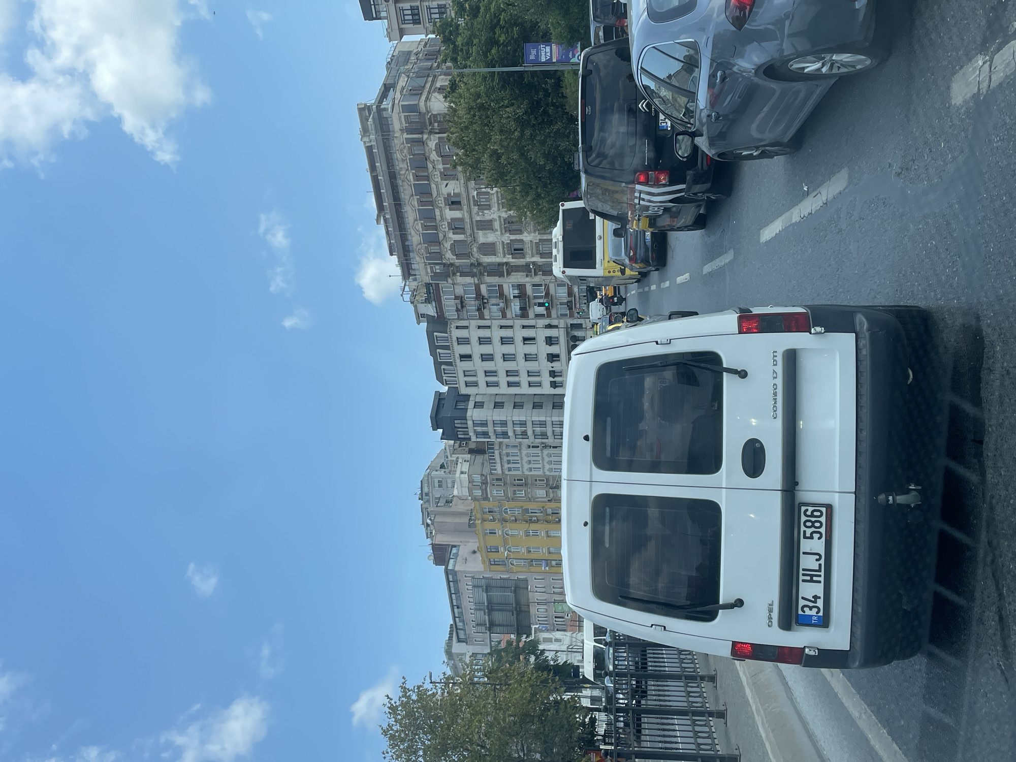 The image depicts a traffic jam on a multi-lane road in Beyoğlu, Turkey. The foreground is dominated by the rear of a white Opel Combo van with a license plate reading "TR 34 HLJ 586." Behind it, several other vehicles are visible, including a black van with "UBER" branding, a gray sedan, and a yellow bus. The road is asphalt with clear lane markings. In the background, a dense cluster of multi-story buildings, characteristic of an urban environment, lines the street. The buildings exhibit a variety of architectural styles and colors, with some featuring ornate detailing. Trees are present along the roadside and interspersed between the buildings. The sky is blue with scattered white clouds. The overall scene suggests a busy urban day with significant traffic congestion.
