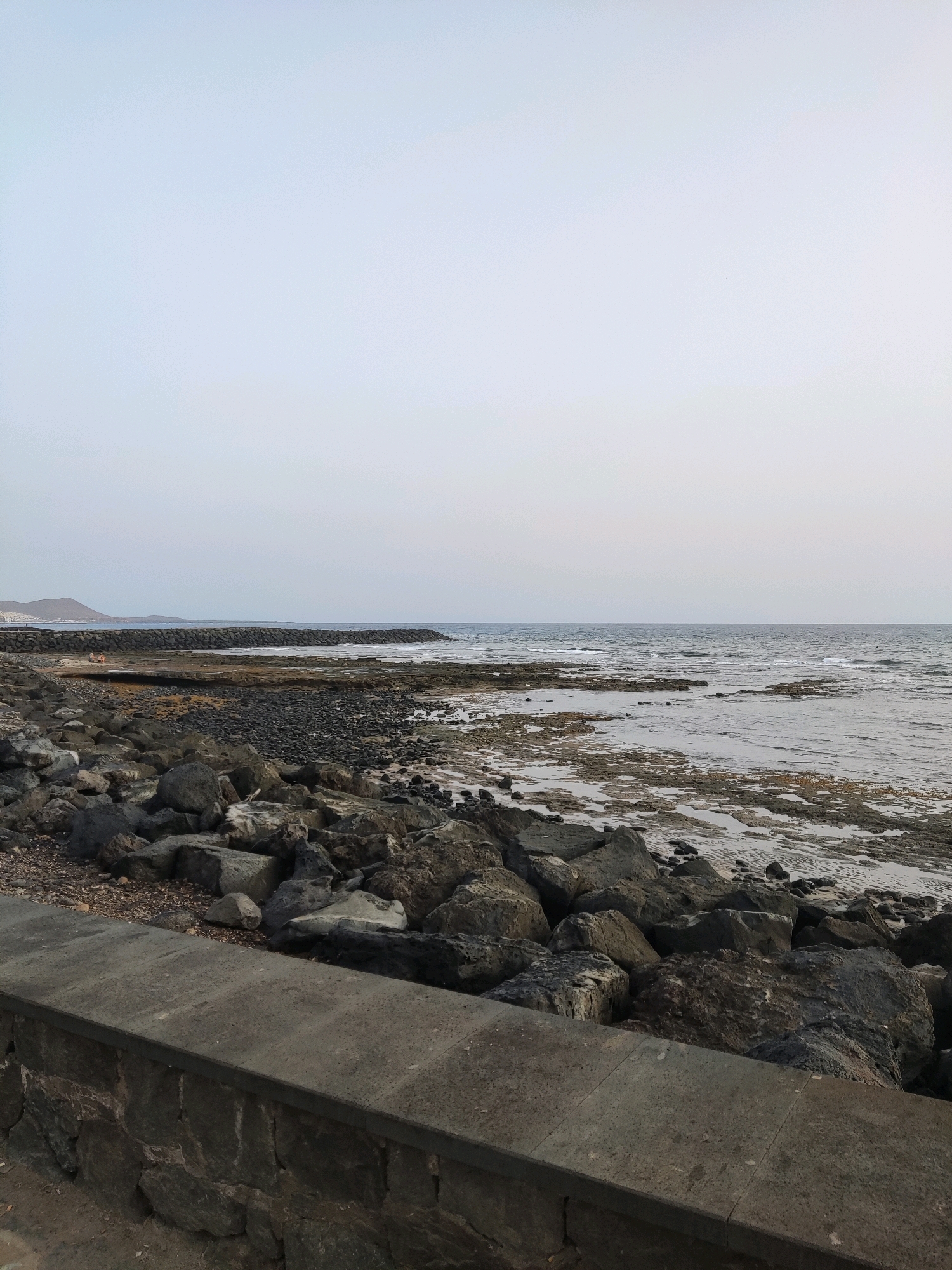 The image shows a rocky coastline with the ocean in the background. The sky is overcast, and the light is muted. In the foreground, there is a low stone wall, and beyond that, a large pile of dark, rough rocks. The ocean is calm, with small waves lapping at the shore. In the distance, a man-made breakwater extends into the water, and further on, a hazy landmass with some buildings can be seen. The area appears to be a public space, possibly a promenade or walking path along the coast. The location is likely Arona, Spain, given the visual cues of a coastal town in a Mediterranean or similar climate. No people are clearly visible and engaged in any actions. The overall scene is one of natural coastal landscape combined with some human-made structures.