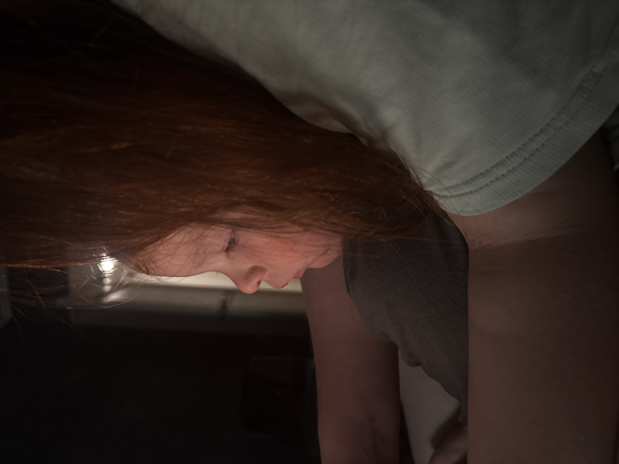 The image is a close-up, side profile of a young person with long, auburn hair. The person is looking down, with their eyes partially closed, suggesting concentration or relaxation. Their features are delicate, with a slightly upturned nose and softly rounded chin. They are wearing a light-colored t-shirt with a ribbed neckline, and a darker grey garment, possibly another shirt or a camisole, is visible underneath.
The lighting is dim, with a strong light source reflecting off the person's hair, creating a halo effect and illuminating some strands. This light also casts shadows, obscuring much of the background, which appears to be dark and indistinct, possibly indicating an indoor setting at night or in a dimly lit room. The overall mood is intimate and quiet, emphasizing the individual's focused or contemplative state. There is no visible text in the image.
