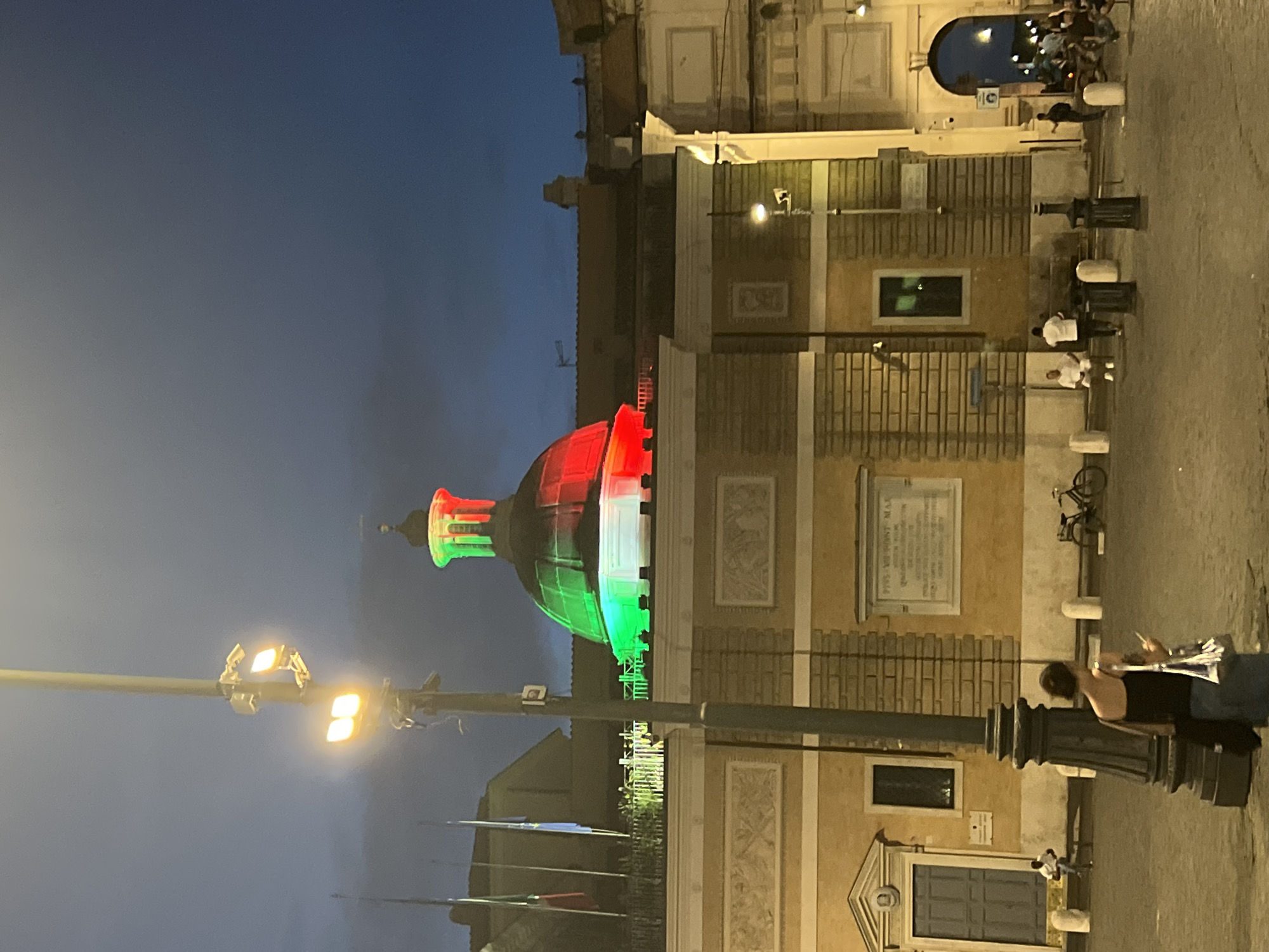 The image captures an evening scene in Rome, Italy, likely near the monument to Victor Emmanuel II due to the Italian flag colors illuminating the dome. The sky is a deep twilight blue. Dominating the upper portion of the image is a building with a prominent dome, which is lit with the colors of the Italian flag: green, white, and red. The building itself is yellow with classical architectural details.
In the foreground, a tall lamppost with multiple lights and surveillance cameras stands prominently. To the left of the lamppost, a woman with dark hair is visible, facing away from the camera, holding a phone and a bag. She is wearing a dark tank top and a blue skirt. In the background, several people are scattered across the paved area. To the left, a person sits on a bench in front of a building entrance. Further back, a bicycle is parked. To the right, a large archway is visible, with more people gathered and walking through it. The ground is paved stone. The overall atmosphere is quiet and serene, with the illuminated dome serving as the focal point.