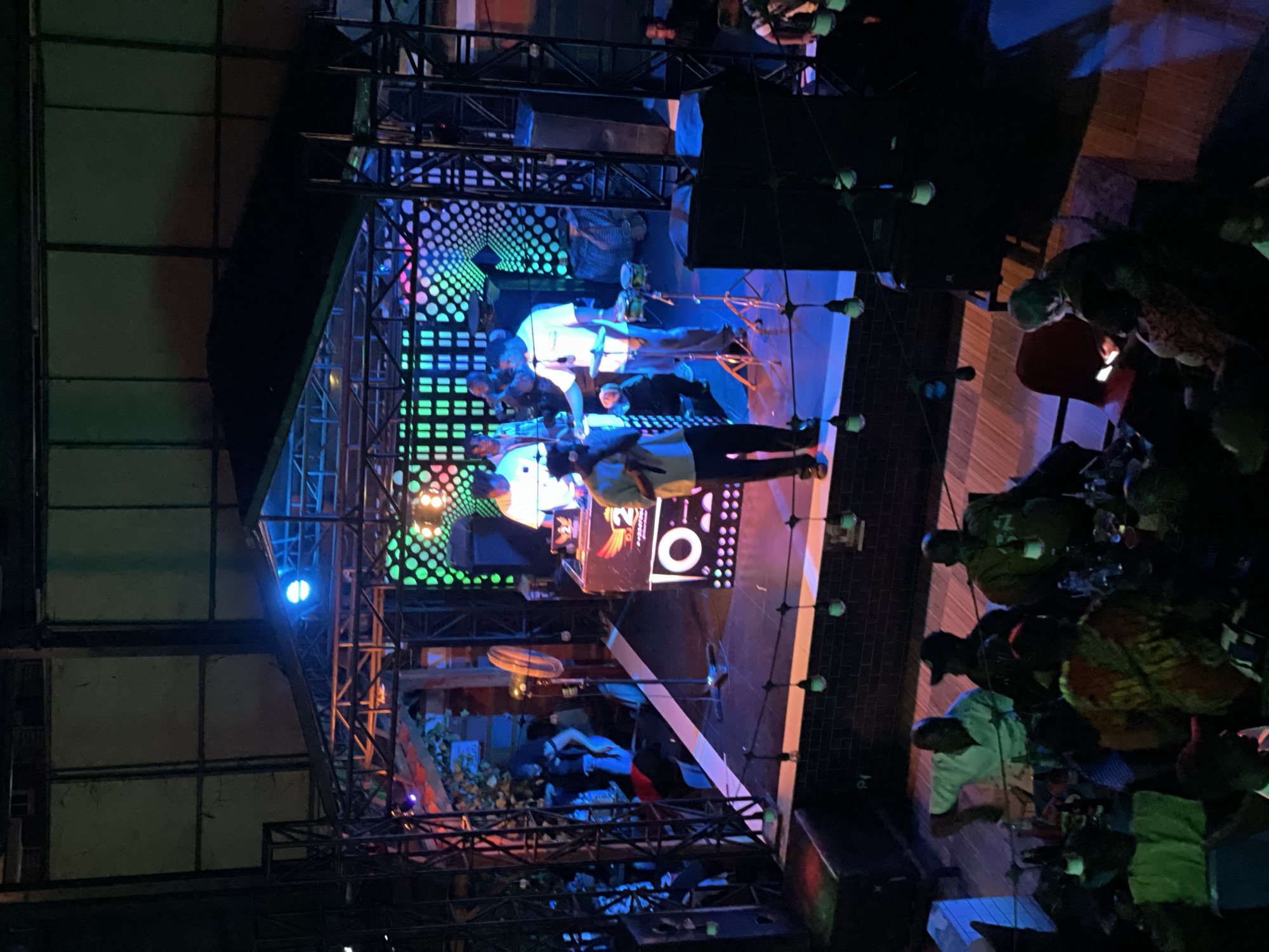 This image captures a lively scene at a venue in Obosi, Nigeria, likely a bar or club, filled with people enjoying a performance. The stage is illuminated with vibrant green and blue lights, featuring a DJ booth and performers. In the foreground, patrons are seated at tables, some with drinks, appearing engaged with the event. The ambiance suggests a social gathering with music and entertainment.
