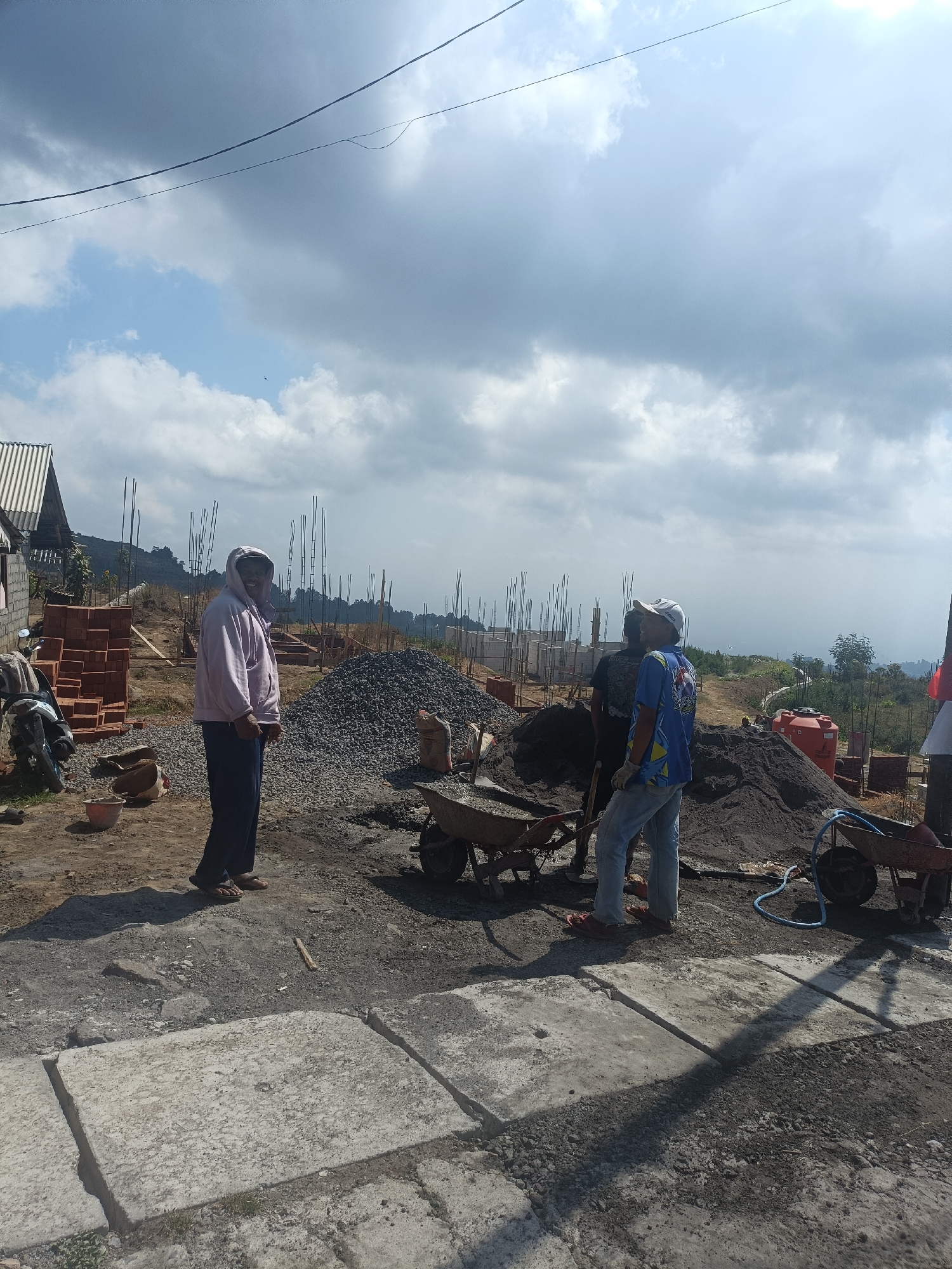 The image depicts an outdoor construction site in Kopeng, Indonesia, under a partly cloudy sky. In the foreground, a person wearing a light-colored hoodie and dark pants stands facing left, with their back partially to the camera. To their right, two other individuals are visible. One, wearing a blue shirt with colorful graphics and a white cap, is facing the person in the hoodie. The second individual, to the left of the person in the blue shirt, is wearing a dark shirt and appears to be looking away.

The immediate area is a dirt and gravel ground, with several piles of construction materials including large mounds of dark grey gravel, heaps of dark sand, and stacks of red bricks. Two wheelbarrows are present, one partially filled with gravel, and another near the sand pile. A red water tank is visible in the mid-ground, and a motorcycle is parked to the left.

In the background, the construction continues with numerous vertical rebar structures protruding from foundations, indicating the early stages of building construction. The landscape slopes upwards behind the site, with sparse vegetation and trees visible on the hills. The overall impression is a busy work environment at a location with an elevated view.