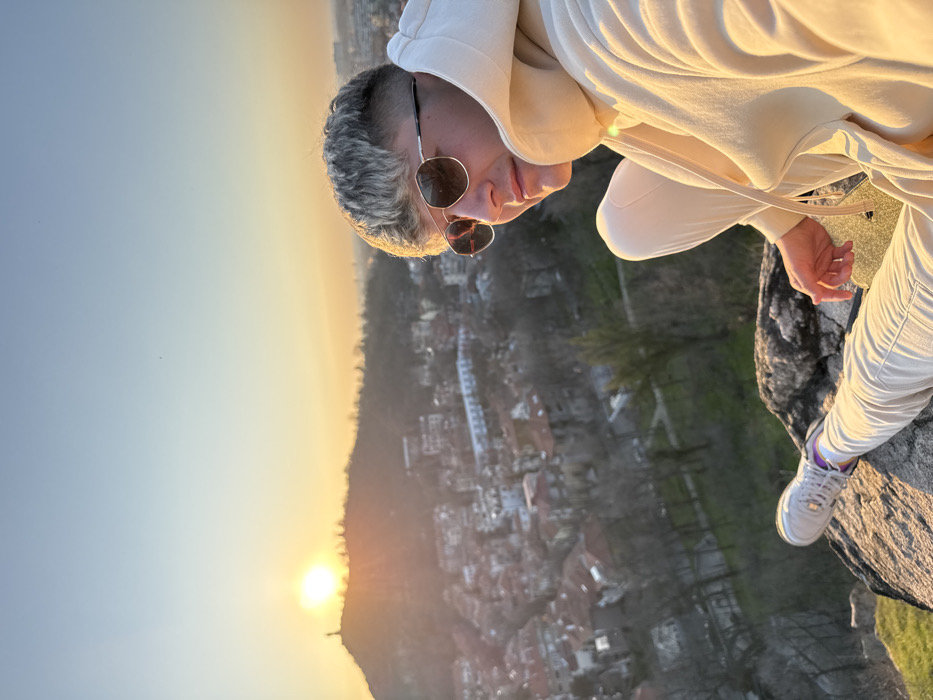 A person is depicted seated on a rock outcrop at sunset, overlooking the city of Plovdiv, Bulgaria. The individual, appearing male, is positioned on a hill, likely Nebet Tepe, with the cityscape spreading out below and to the west-southwest. The sun is setting behind Bunardzhika Hill (Hill of Liberators), directly silhouetting the Alyosha Monument which stands atop it.
The person has short, light-colored hair and wears dark, reflective aviator-style sunglasses. They are dressed in a light beige hooded sweatshirt and matching sweatpants, paired with white sneakers that feature purple accents. The individual is seated with their knees bent, their left leg extended forward, and their right arm resting on their right leg. A small, gold-colored, glittery object is partially visible near their right hand.
The immediate foreground features large, rugged rock formations illuminated by the low sun, with patches of green vegetation. The midground presents a sprawling urban environment with numerous residential buildings, many displaying orange-tiled roofs, interspersed with bare trees and areas of parkland. The sky above is clear, transitioning from a bright orange-yellow at the horizon, where the sun is prominently visible, to a pale blue overhead.
