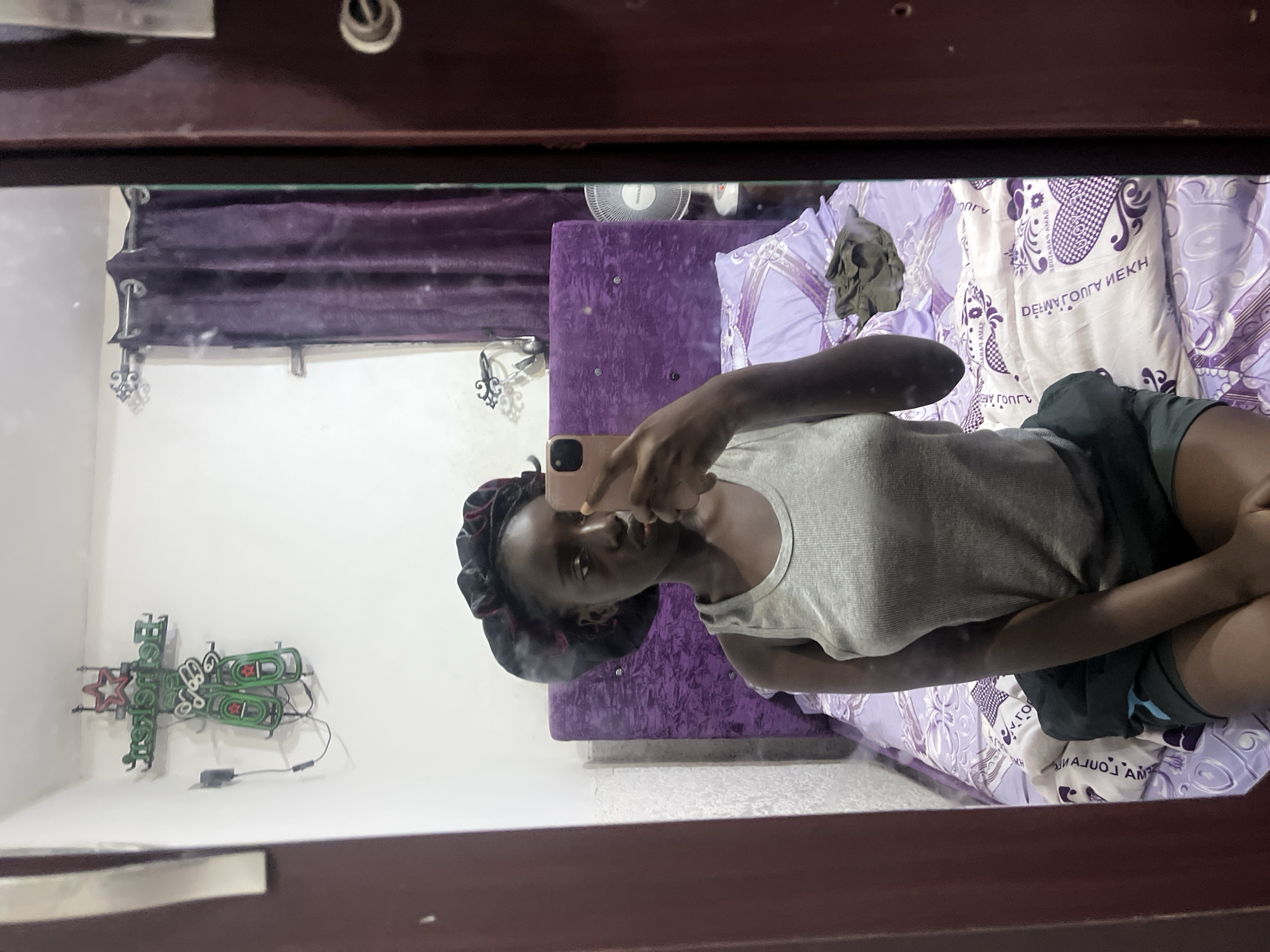 A medium shot captures a person in a mirror, taking a selfie. The person, likely female, wears a satin hair cap and a light-colored tank top. She is positioned on a bed with patterned white and purple sheets. Behind her is a purple headboard. The reflection reveals purple curtains on the wall, and a "Heineken" neon sign is mounted on the wall to the left. A ceiling fan can be seen. The setting appears to be a bedroom. Without additional information, it's impossible to verify the location in Abuja, Nigeria.