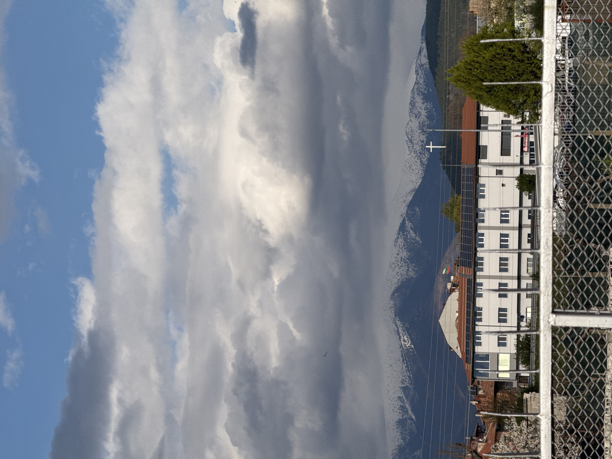 The image shows a view in Blagoevgrad, Bulgaria, looking towards a mountain range partially obscured by clouds. A building is visible with solar panels on its roof, and a white cross stands on a hill in front of the mountain. The image is taken through a chain-link fence. The sky is partly blue with scattered white and gray clouds. A bird is visible in the sky.