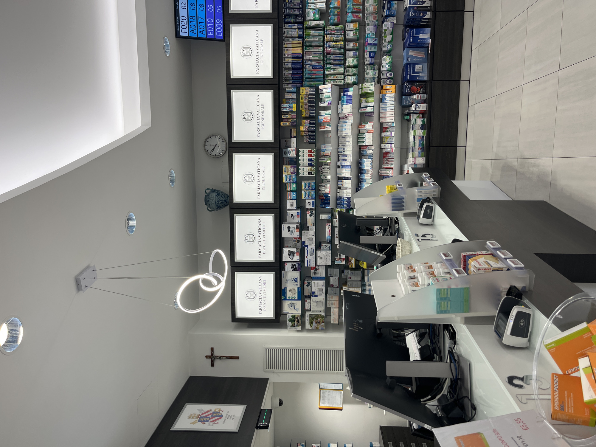 This image depicts the interior of a pharmacy, likely located in Città del Vaticano, Vatican City. The scene is brightly lit, with modern fluorescent and pendant lighting fixtures visible on the ceiling. A counter area dominates the foreground, featuring computer monitors, a card reader, and various pharmaceutical products. Shelving units are packed with a wide array of health and personal care items, including medications, toothbrushes, and dental hygiene products. Signs indicating "Farmacia Vaticana" are prominently displayed. A wall features framed artwork, including what appears to be a religious crest and a crucifix. The overall impression is that of a clean, organized, and well-stocked retail environment for health-related goods.