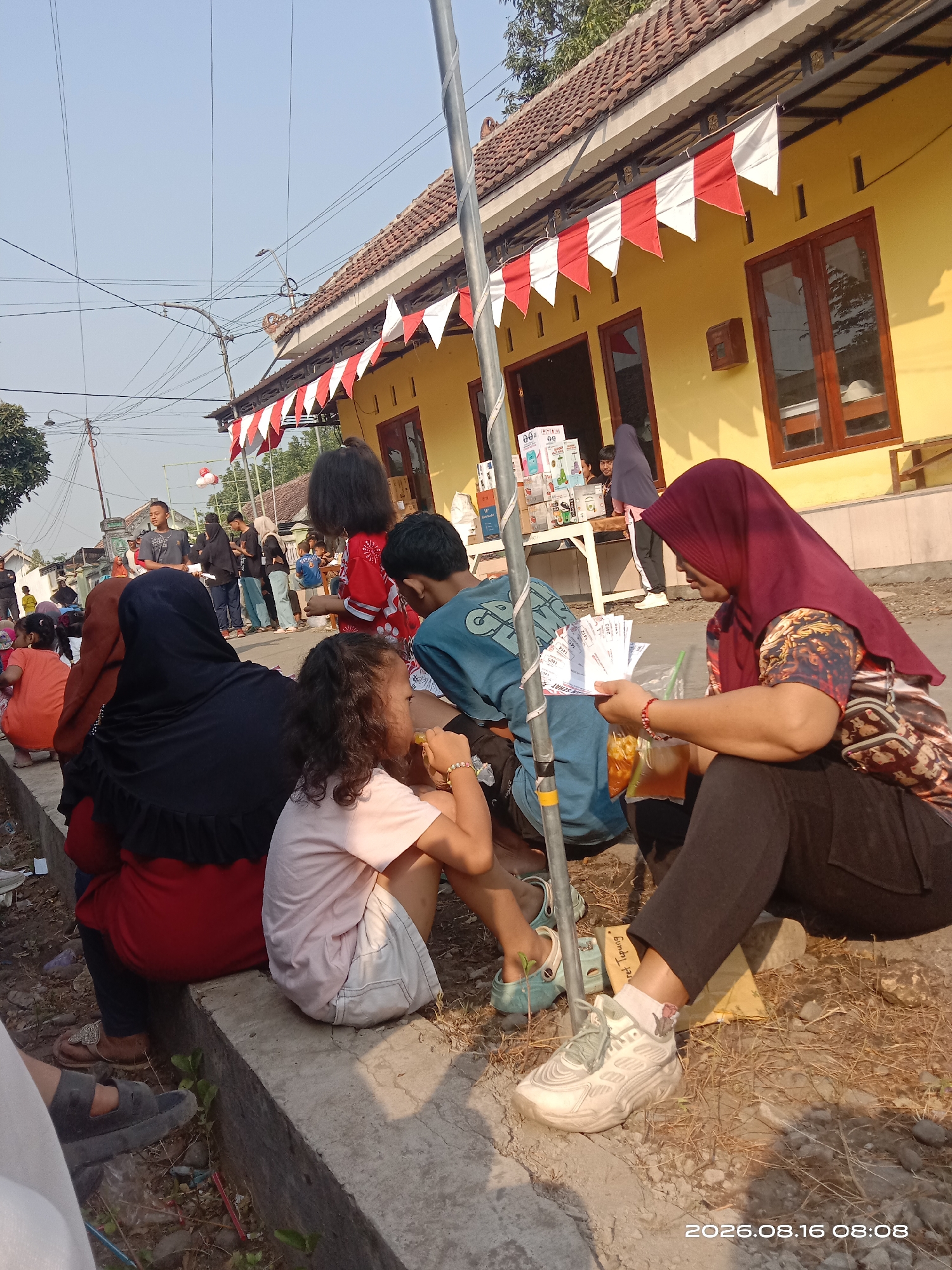 In Banyuarang, Indonesia, a gathering of people, including children and adults, is taking place on a street in front of a yellow building adorned with red and white flags. Several individuals are seated on a concrete barrier, while others stand or walk. A woman in a maroon hijab is holding a bag of snacks and some papers, appearing to be in the process of a transaction. Nearby, a child in a pink shirt and white shorts is also seated, looking towards the woman. Other people are seen in the background, some interacting with a vendor's stall that displays various products. The scene suggests a local event or market day, with a casual and community-oriented atmosphere.