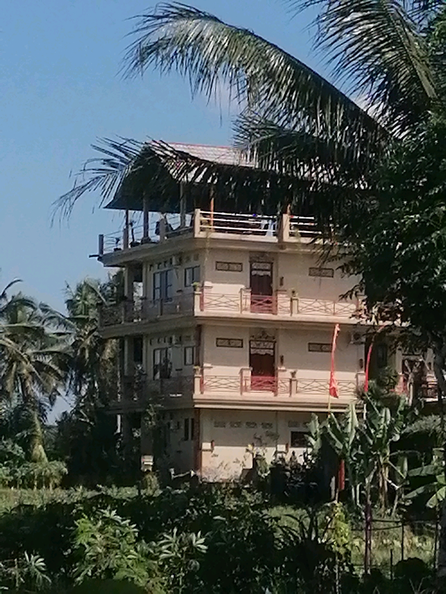 This is a medium shot, eye-level, horizontal, full-color photograph of a multi-story building resembling a guesthouse or hotel, situated in the town of Ubud, Bali, Indonesia. The building is light beige with white trim and balconies, and has a thatched roof structure on its uppermost level. There are several balconies with decorative red railings, and windows with dark frames. The building is partially obscured by lush green foliage, including palm trees and other tropical plants. The sky is a clear, pale blue. The scene suggests a tropical, possibly resort-like setting, consistent with the environment of Ubud.