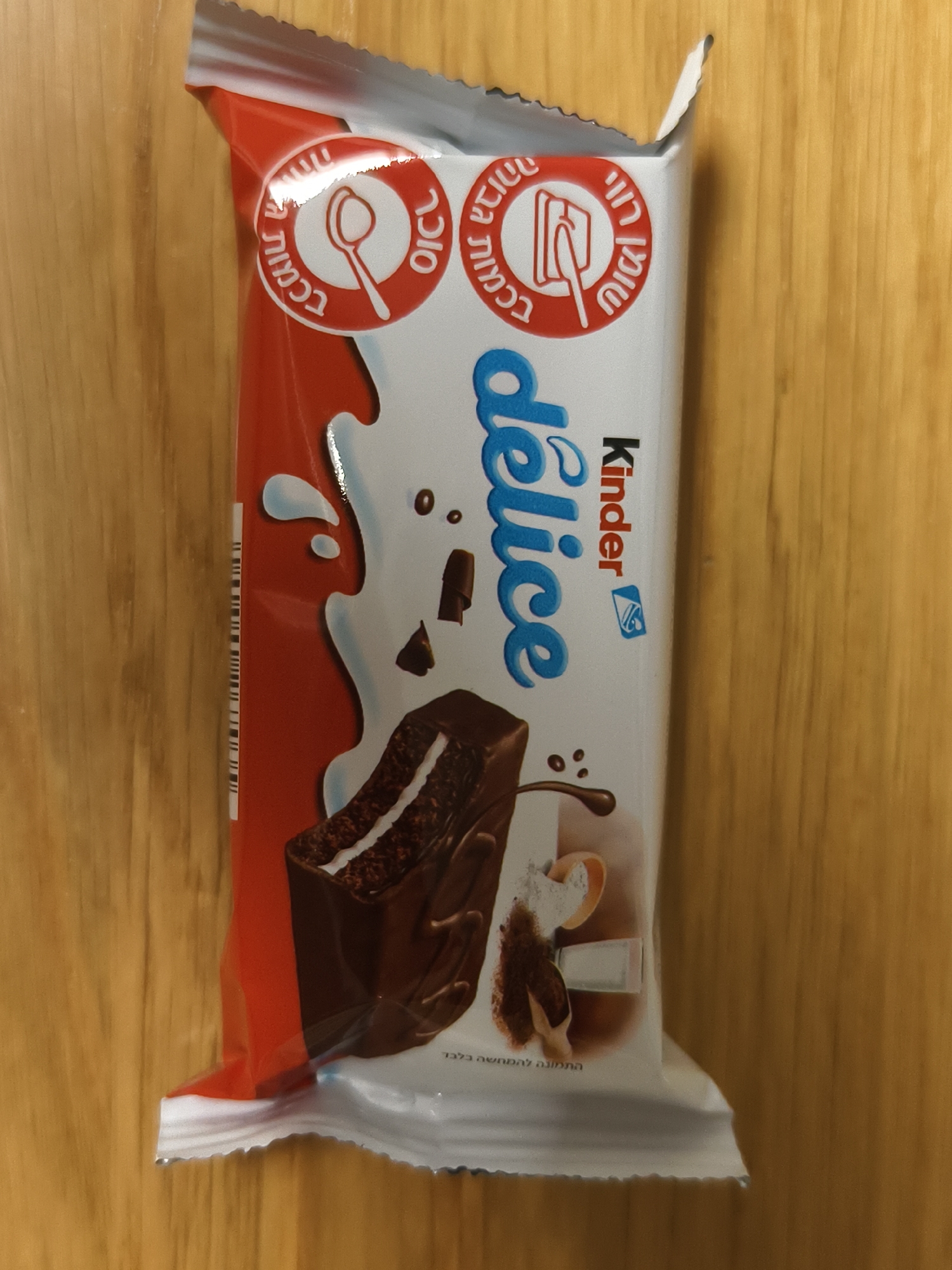 A package of Kinder Delice is placed on a light brown wooden surface. The package is red and white and features an image of a chocolate dessert. The text on the package includes "Kinder Delice" in blue lettering and Hebrew text. Petah Tikva, Israel is not mentioned in the image.