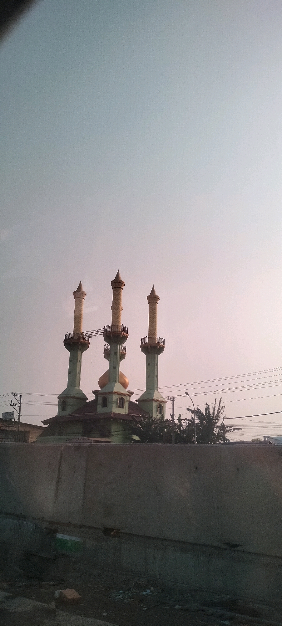 A mosque with three tall, slender minarets stands against a pale sky. The minarets are green at the base, transitioning to a textured yellow towards their conical tops, each topped with a golden finial. A golden dome sits atop the mosque's main structure, which is also primarily green. The mosque is elevated and appears to be part of a complex that includes other buildings, greenery, and utility poles with electrical wires. In the foreground, a stark concrete barrier runs horizontally across the lower portion of the image, obscuring the base of the mosque. The sky exhibits a soft gradient of light blue at the top, fading into a pinkish hue towards the horizon, suggesting either dawn or dusk. The overall scene is calm and depicts a religious building in an urban or semi-urban environment. This mosque is likely the Masjid Agung Jawa Tengah in Semarang, Indonesia, due to its distinctive triple minarets and golden dome.