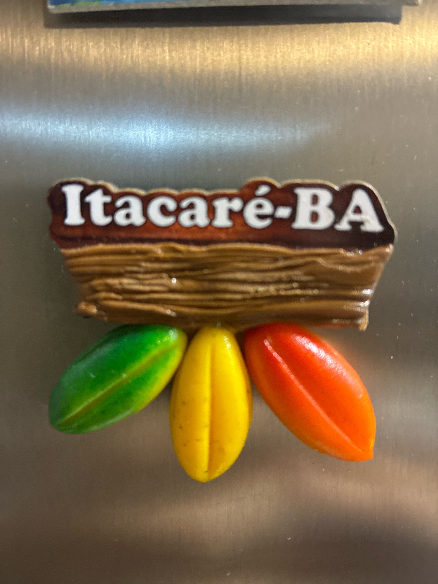 The image shows a close-up of a refrigerator magnet. The magnet has a brown, wood-like texture at the top, with the text "Itacaré-BA" printed in white and outlined in brown. Below this, three colorful, pod-like shapes are arranged in a cluster: a green one on the left, a yellow one in the middle, and an orange one on the right. The background is a metallic surface, likely the side of a refrigerator, with visible horizontal brush strokes and some subtle reflections. There are no people, actions, or interactions depicted. The location is not Curitiba, Brazil, but Itacaré, Bahia, Brazil, as indicated by the text on the magnet.