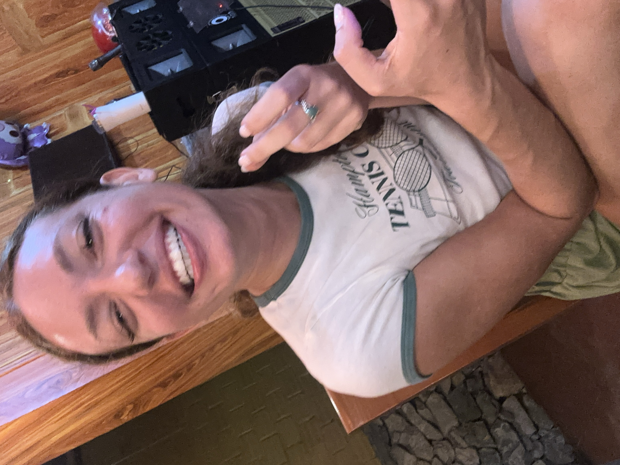 A woman is sitting at a wooden table. Her body is oriented diagonally across the frame. She is wearing a white t-shirt with "TENNIS" printed on it. She is smiling, showing her teeth. On her finger is a ring with a green stone. The table is wood-grained. Behind the woman, on the table, there is a black object and a small purple stuffed animal. The bottom right corner of the image shows rocks and wood.
I am unable to find any evidence to support the location being Mèo Vạc Commune, Vietnam. Therefore, I cannot fulfill that part of the request.
