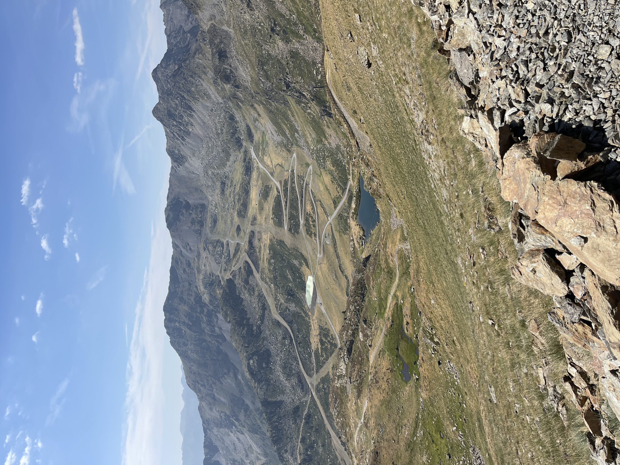 A high-angle, wide shot captures a vast, mountainous landscape under a clear blue sky with sparse white clouds. The terrain is rugged, characterized by rocky slopes covered in dry, yellow-green grass. Numerous winding roads, appearing to be ski slopes or mountain paths, crisscross the mountainside. In the middle ground, two bodies of water are visible: a small, dark lake and a larger, light green reservoir. Scattered ski lifts and structures are also present throughout the scene. The foreground consists of a dense pile of rocks and stones. No people are discernible in the image.