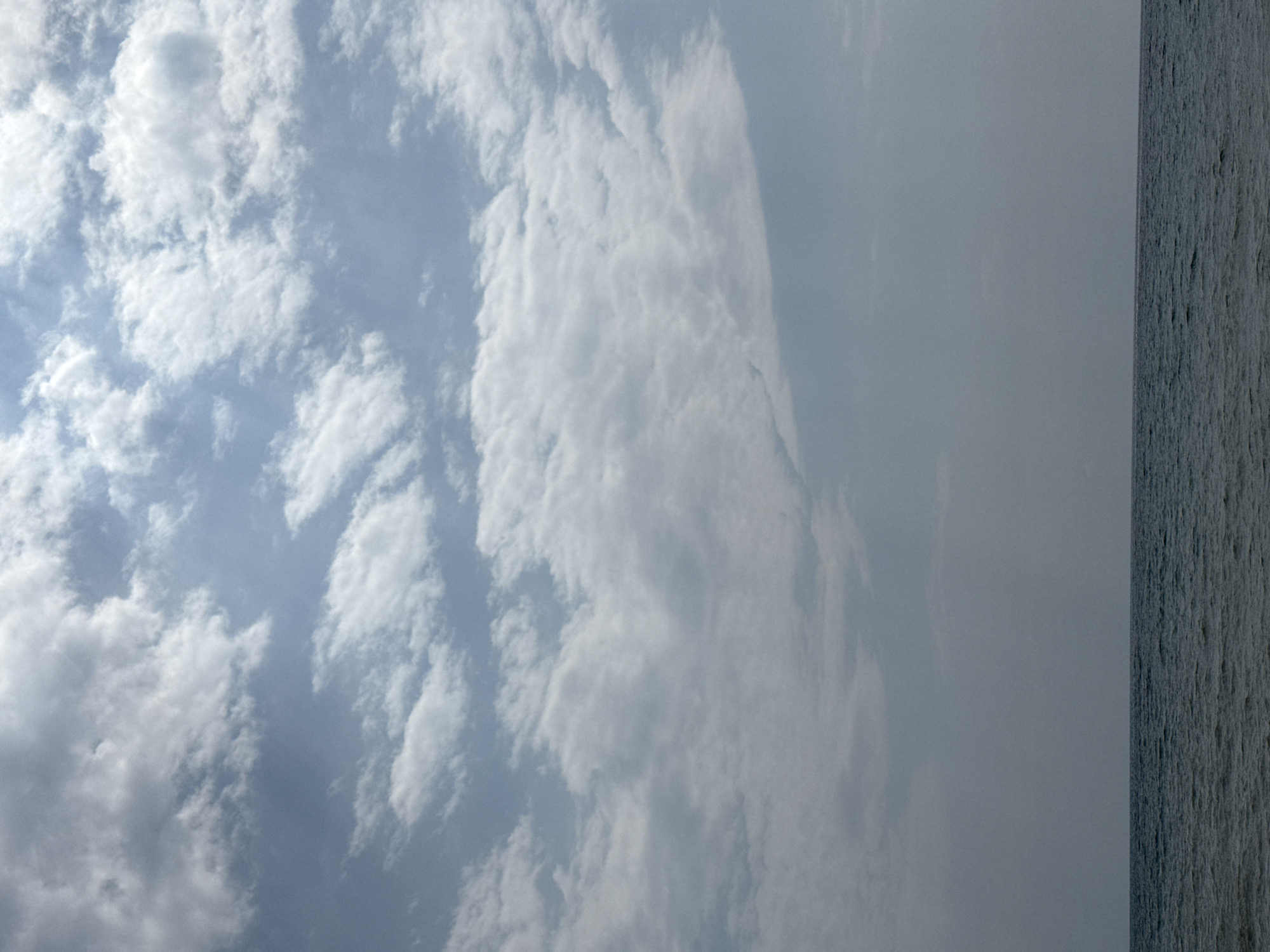 The image depicts a wide, horizontal seascape under a sky filled with scattered clouds. The horizon line is clearly defined, separating the deep blue-grey water from the pale blue sky. The clouds are varied, with some appearing wispy and translucent, while others are more dense and textured, casting subtle shadows. The water exhibits a gentle ripple, suggesting a calm, though not entirely still, body of water. The overall impression is one of open space and natural beauty, with no man-made structures or living beings visible in the frame. The scene is likely a coastal or lakeside view. Given the provided context, this view is situated in the city of Sary-Oy, Kyrgyzstan, implying it is a view of Issyk-Kul Lake.
