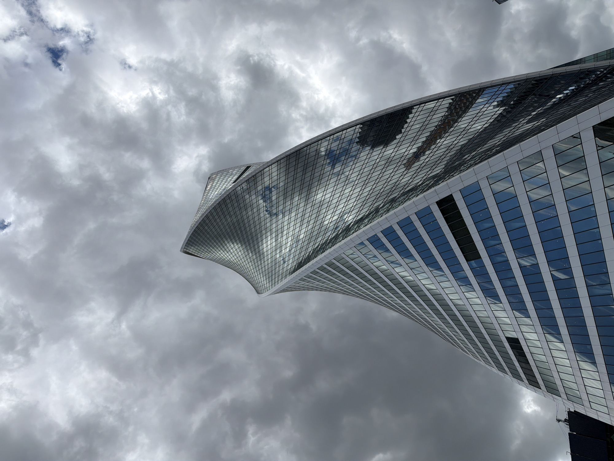 The image captures a low-angle view of the Evolution Tower in Moscow, Russia. The tower has a distinct twisted design, with a glass facade reflecting the cloudy sky. The sky is predominantly overcast, with varying shades of gray and hints of blue. The lower section of the tower is visible, showing the building's construction and window patterns. The overall scene emphasizes the architectural design against the backdrop of the sky.