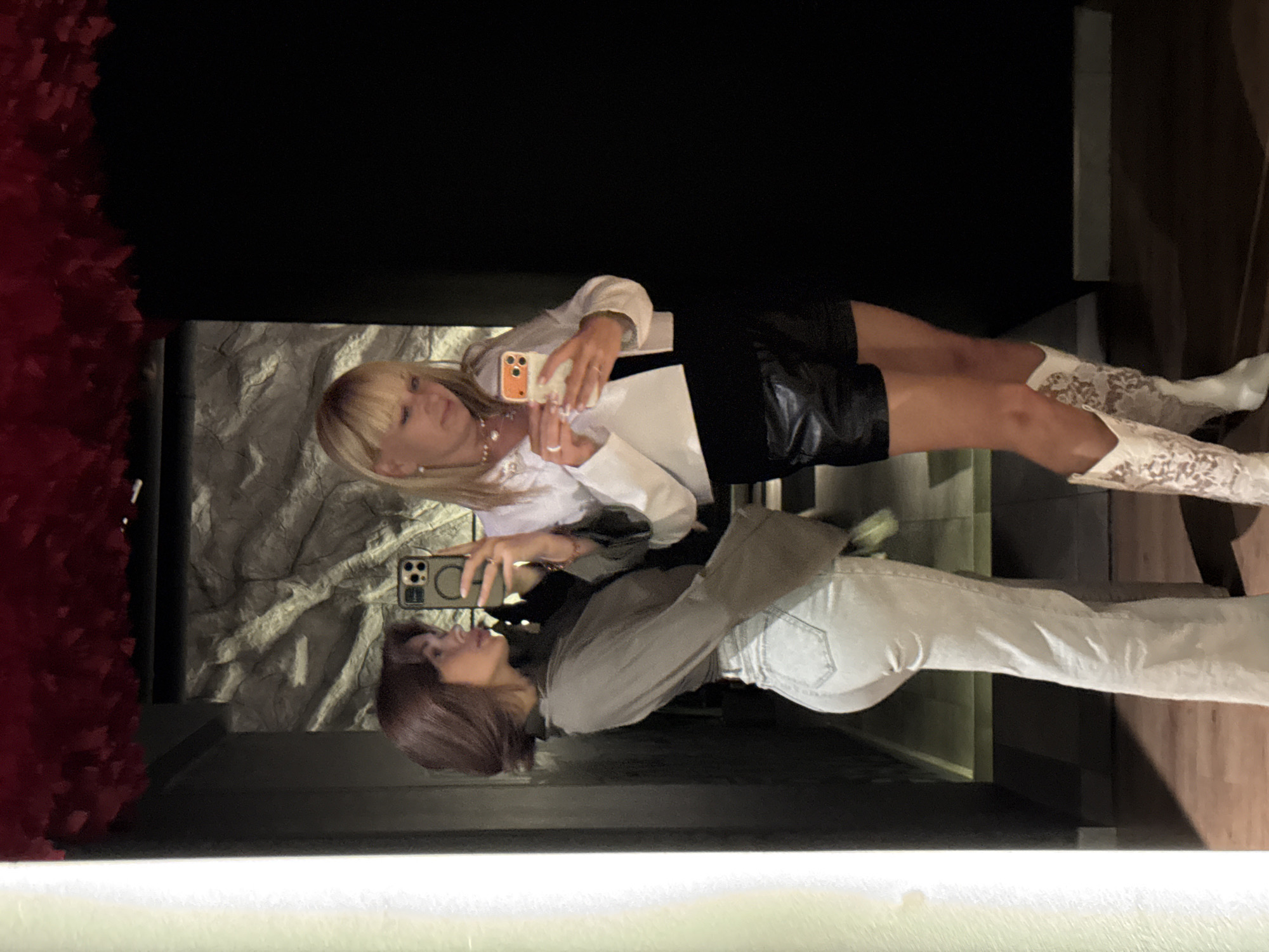 Two women are taking photos of themselves in a mirror. The woman on the right wears a white long-sleeved shirt with a black skirt and white lace boots. She holds an orange smartphone. The woman on the left wears a grey long-sleeved shirt and light blue jeans. She holds a green smartphone. A wall covered in red fabric is on the left. The walls of the room are dark, and a stone-like wall is visible in the mirror. The location is Chaikovsky, Russia.