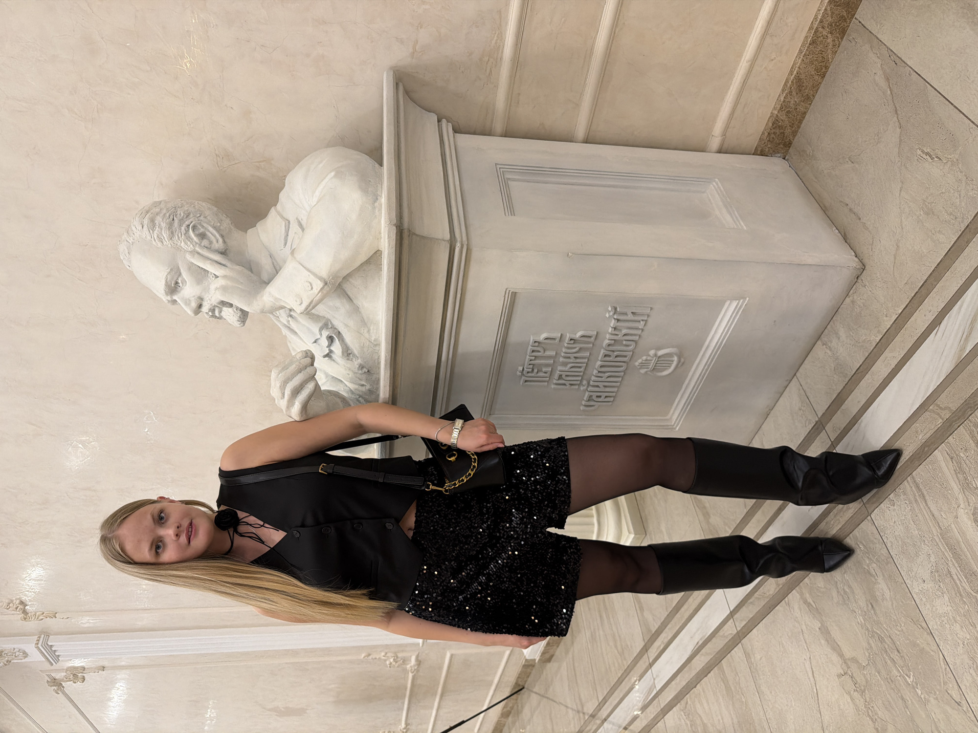 A young blonde woman is standing next to a bust of Tchaikovsky in Izhevsk, Russia. She is wearing a black, sparkling dress, black tights, and black boots. She has a black purse slung over her shoulder. The bust of Tchaikovsky is white and sits on a white pedestal. The background is a beige wall and floor with white trim.