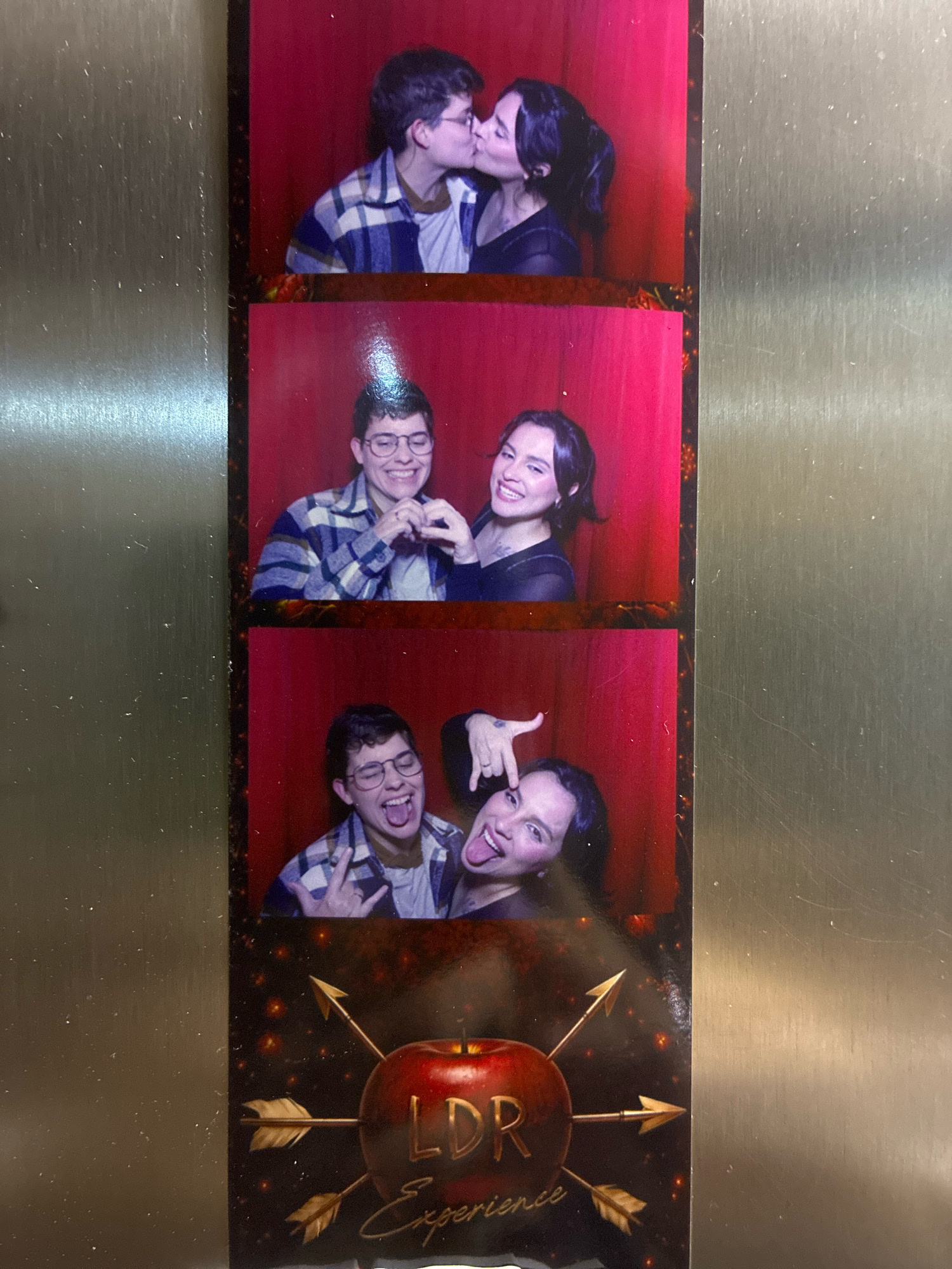 The image is a vertical, three-panel photo booth strip with a metallic background bordering the right side. Each panel features two individuals against a red backdrop. The top panel shows a person with short, dark hair and glasses kissing another person with dark hair pulled back in a ponytail. Both are wearing casual clothing, with one in a plaid shirt. The middle panel shows the same two people, both smiling. The person with glasses is wearing a plaid shirt, and the other person has dark hair and is wearing a dark top. The bottom panel shows the two people again, this time making playful faces. The person with glasses is sticking their tongue out and making a rock-on hand gesture, while the other person has their tongue sticking out and is pointing at their eye. Below the photo strip is a graphic featuring a red apple pierced by three gold arrows, with the text "LDR Experience" in gold lettering. The overall scene depicts a fun, lighthearted interaction between two individuals in a photo booth. This photo booth strip is located at the "LDR Experience" event, which is being held at the Expo Renault Barigui in Curitiba, Brazil.