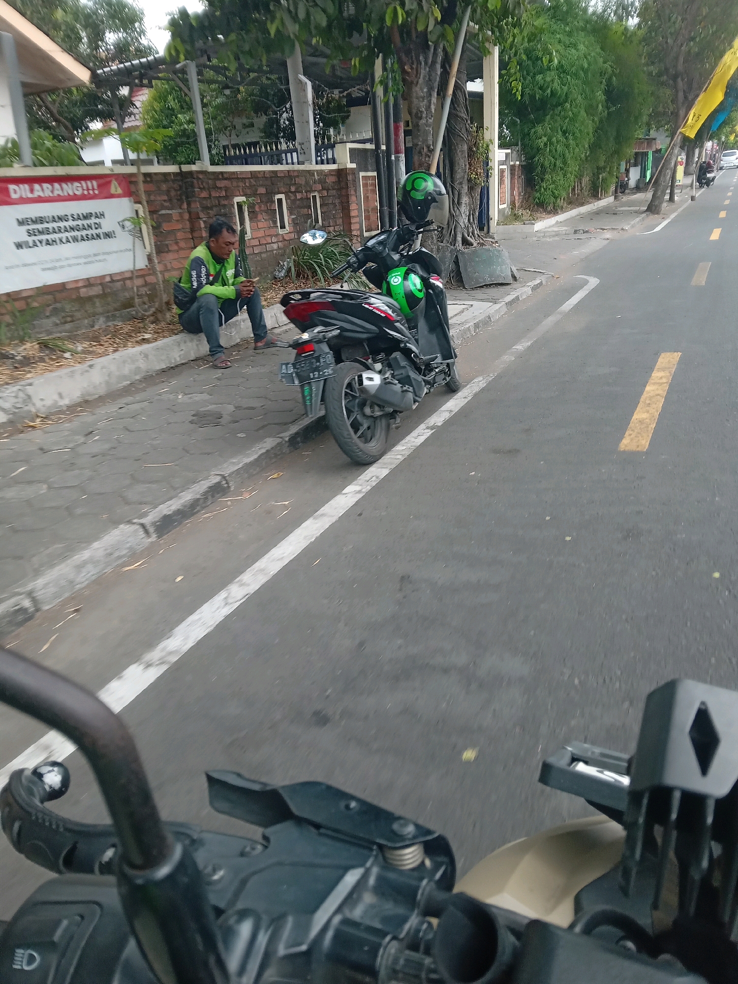 A man wearing a green jacket with "Gojek" text and dark pants sits on a gray concrete curb beside a brick wall. He is holding a smartphone and appears to be looking at it. A black motorcycle with a green helmet on its seat is parked behind him. A white sign with red lettering that reads "DILARANG!!!" (Forbidden!!!) is attached to the brick wall to the left of the man. The sign also has text below stating "MEMBUANG SAMPAH SEMBARANGAN DI WILAYAH KAWASAN INI!" (Dumping garbage indiscriminately in this area is prohibited!). The scene is on a street in Yogyakarta, Indonesia, with a paved road and sidewalk. Trees and buildings are visible in the background. The image is taken from the perspective of someone on a motorcycle, with handlebars and a camera mount visible in the foreground.