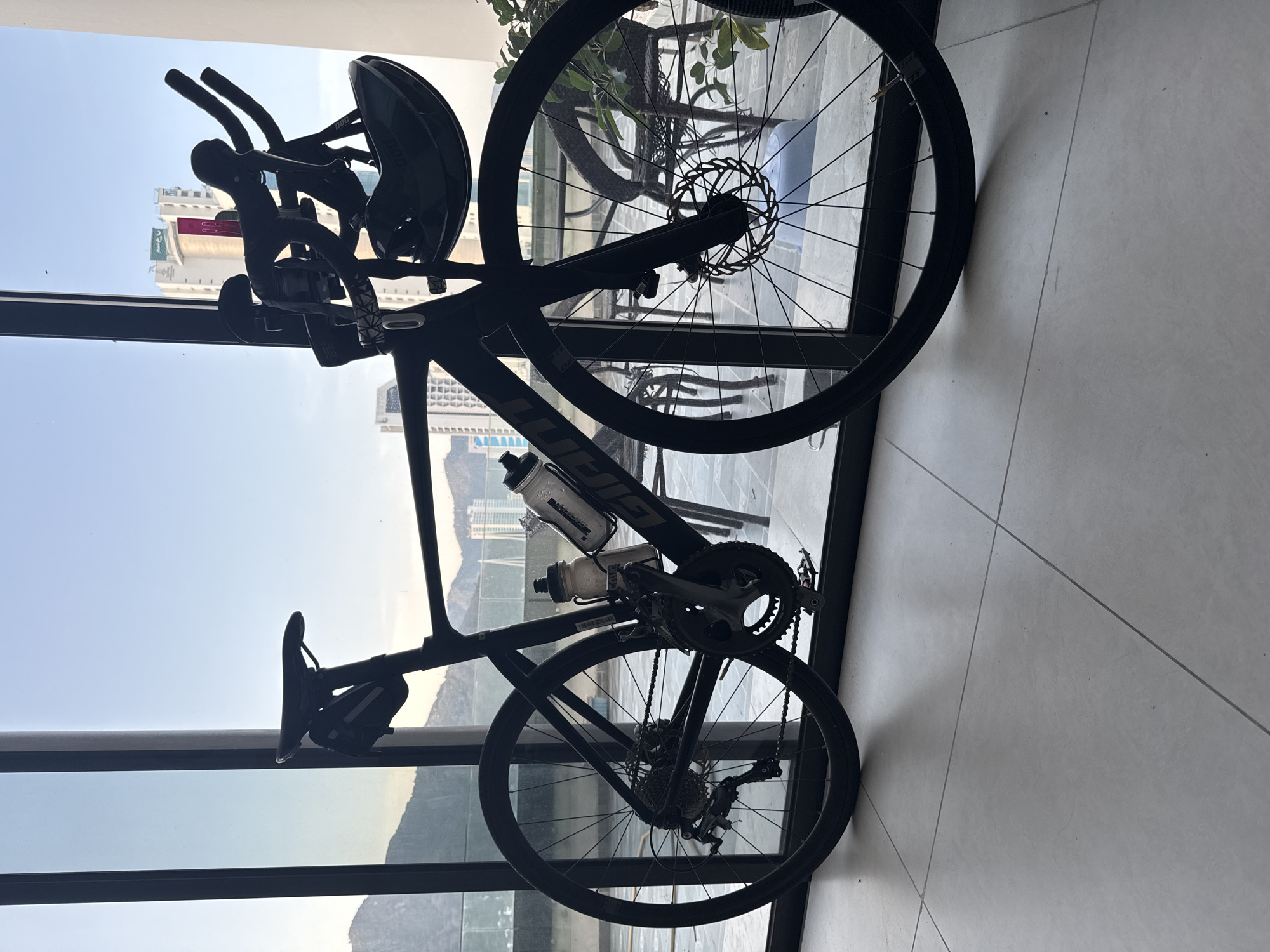 A matte black Giant road bicycle, equipped with aero handlebars, a black helmet, a seat bag, and two white water bottles, stands indoors on a light grey tiled floor. The bicycle's disc brakes, rear derailleur, cassette, and clipless pedals are visible. The scene is viewed through large, dark-framed, floor-to-ceiling windows that separate the interior from an outdoor terrace. Beyond the terrace, a panoramic view of Bac Nha Trang Ward, Vietnam, is visible under a clear blue sky. In the distant background to the left, mountains are silhouetted against the horizon. In the mid-ground, modern high-rise buildings dominate, including one with a distinctive red vertical element and "A&EM" and "Green World Hotel" signage. On the outdoor terrace directly outside the window, dark woven outdoor chairs and tables are arranged, along with a light blue plastic container and green foliage from a potted plant. The terrace floor appears to be paved.