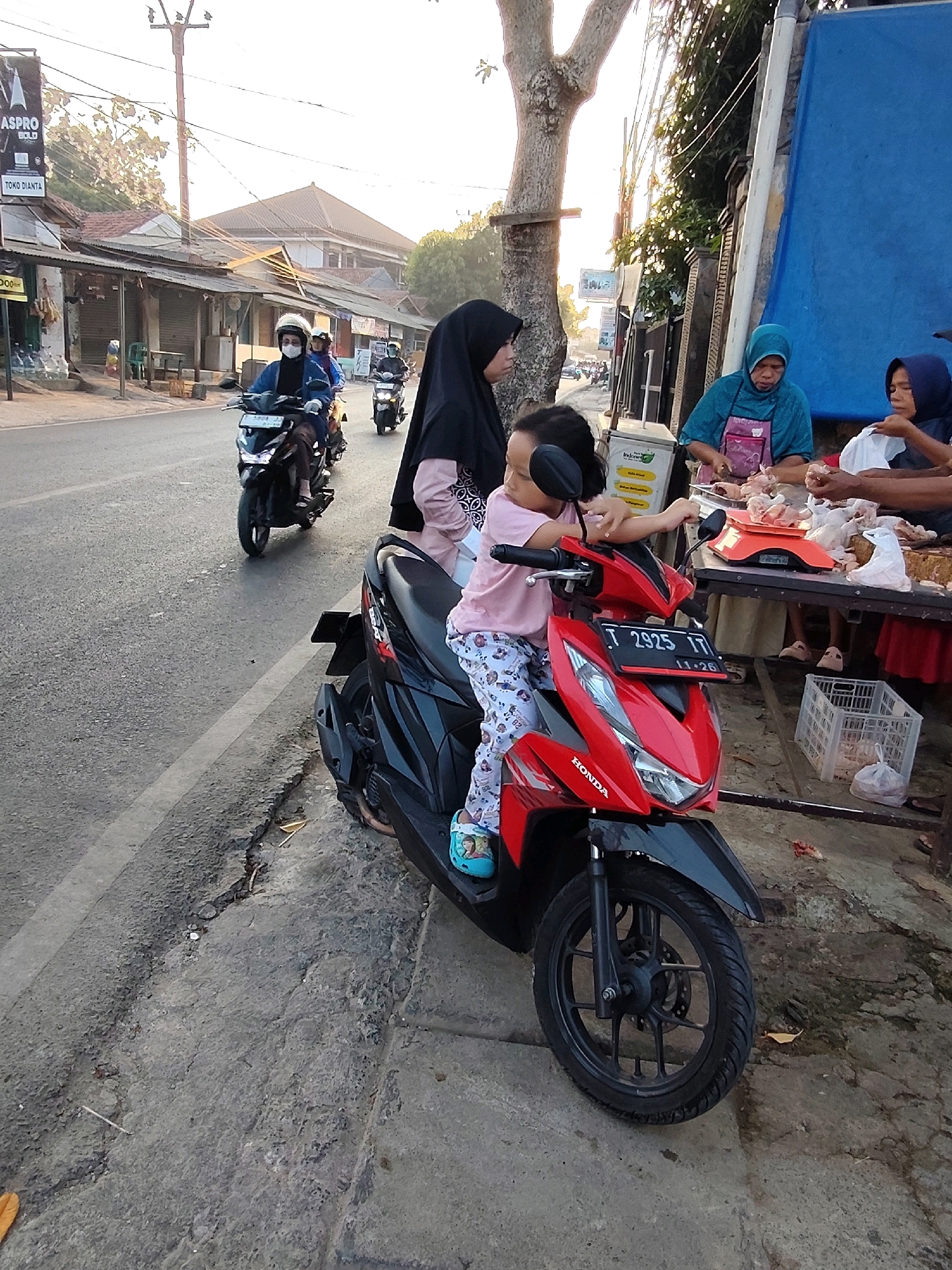 In the city of Purwakarta, Indonesia, an outdoor scene unfolds at a roadside market. The sun casts a warm glow on the asphalt road and surrounding area. In the foreground, a child in a pink shirt and patterned leggings sits on a red Honda motorcycle, the child's left arm resting on the handlebars. The motorcycle is parked on the side of the road, facing away from the camera.
Behind the motorcycle, a woman wearing a black hijab and pink shirt is seated, also appearing to be on the motorcycle. To the right of the motorcycle, a market stall is set up with a table laden with chickens. Two women are engaged in activities at the stall; one, wearing a teal hijab and apron, is preparing chickens, while another, in a dark blue hijab, holds a plastic bag. A red digital scale is visible on the table, with several chickens on it. A white plastic crate sits on the ground near the table, containing plastic bags.
In the background, several motorcycles are seen on the road. On the left side of the road, a row of shops with corrugated metal roofs lines the street. A sign with "ASPRO" and "TOKO DIANTA" is visible on one of the buildings. The environment includes a large tree to the left of the market stall and a blue tarp hanging behind it. The overall scene depicts a typical day at a local Indonesian market.