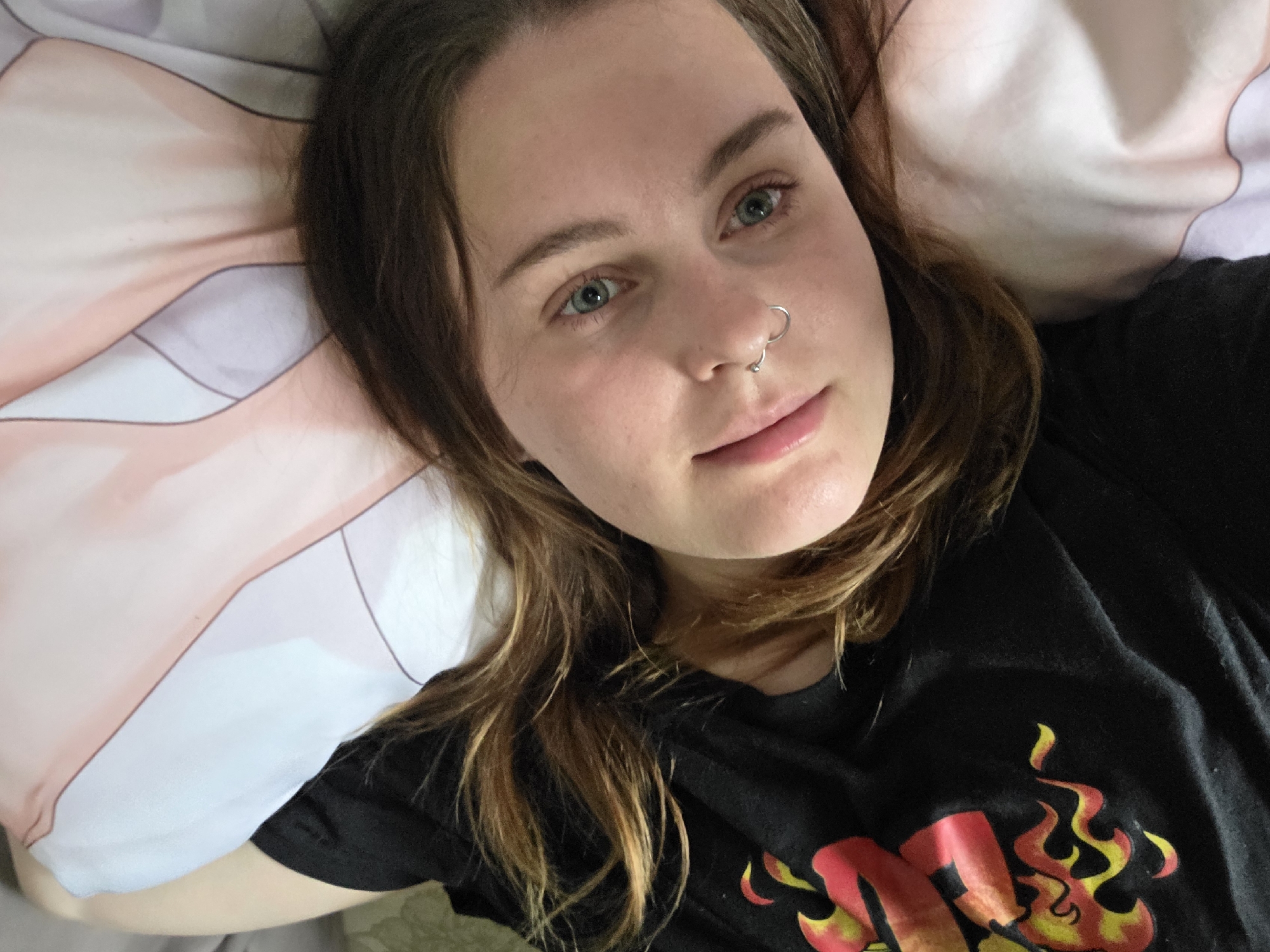 A person with fair skin and light brown hair with blonde streaks is depicted lying on their back, facing upward, and looking directly into the camera with a subtle, closed-mouth smile. The individual has greenish-blue eyes and a small silver hoop piercing through the nasal septum. They are wearing a black short-sleeved t-shirt with a graphic design on the left chest area, consisting of red and yellow flame-like patterns. Their hair is fanned out on a pillow behind their head. The pillow features a pattern of light pink and white geometric or stylized shapes. A cream-colored, textured surface is visible along the far left edge of the frame. The scene is illuminated by soft, diffused lighting. The location is Unknown, Unknown.
