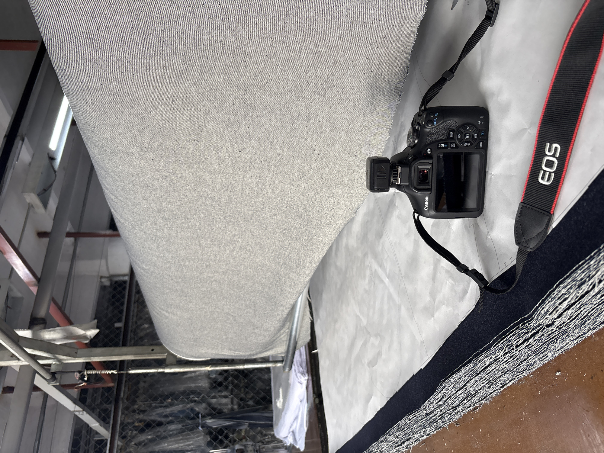 The image shows a large roll of gray fabric, presumably in a textile factory or warehouse. A Canon EOS camera with a flash attachment sits on top of the fabric. The camera has a black strap with the "EOS" logo in white. In the background, there is industrial machinery, including metal supports and what looks like a chain-link fence. There are also some pieces of white fabric visible in the background. The photograph appears to be taken in a factory setting, and the presence of the camera suggests someone is documenting the textile process or inventory. The location is unknown.