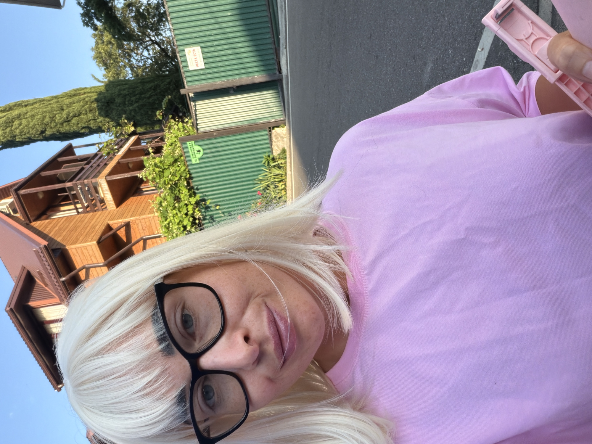 A fair-skinned woman with blonde hair and glasses takes a selfie outdoors on a street in Sochi, Russia. She wears a pink t-shirt and holds a pink object, possibly a stapler, in her right hand. Behind her, a wooden house with balconies and a green fence are visible under a clear blue sky. The image captures a sunny day with warm lighting.