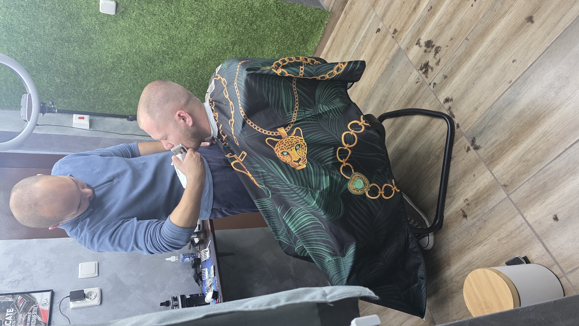 The image, originally rotated 90 degrees to the left, shows an indoor grooming session. Two bald men are the primary subjects. One man, wearing a dark blue long-sleeved shirt, is actively using a white electric shaver to trim the head or neck of the other man. The man receiving the grooming is covered by a distinctive black cape featuring a pattern of gold chains, green palm leaves, and a large gold leopard head pendant with green eyes.
The setting appears to be a modern barbershop or a dedicated grooming area. The floor is covered with light brown wooden planks or tiles, some with small dark spots that resemble hair trimmings. One wall has a textured gray finish, displaying a power outlet with a black charger plugged in, and a partially visible framed item with text like "CATE", "THE COURSE OF", and "FUNDAMENTALS". Another section of the wall or a backdrop is covered in artificial green grass. A ring light on a stand is visible on the left side of the frame, suggesting attention to lighting, possibly for detailed work or content creation. A small black chair frame and a light-colored cylindrical trash can with a wooden lid are also visible on the floor. Both individuals appear focused on the grooming activity. The scene is likely taking place during the daytime in Redina, Bulgaria.