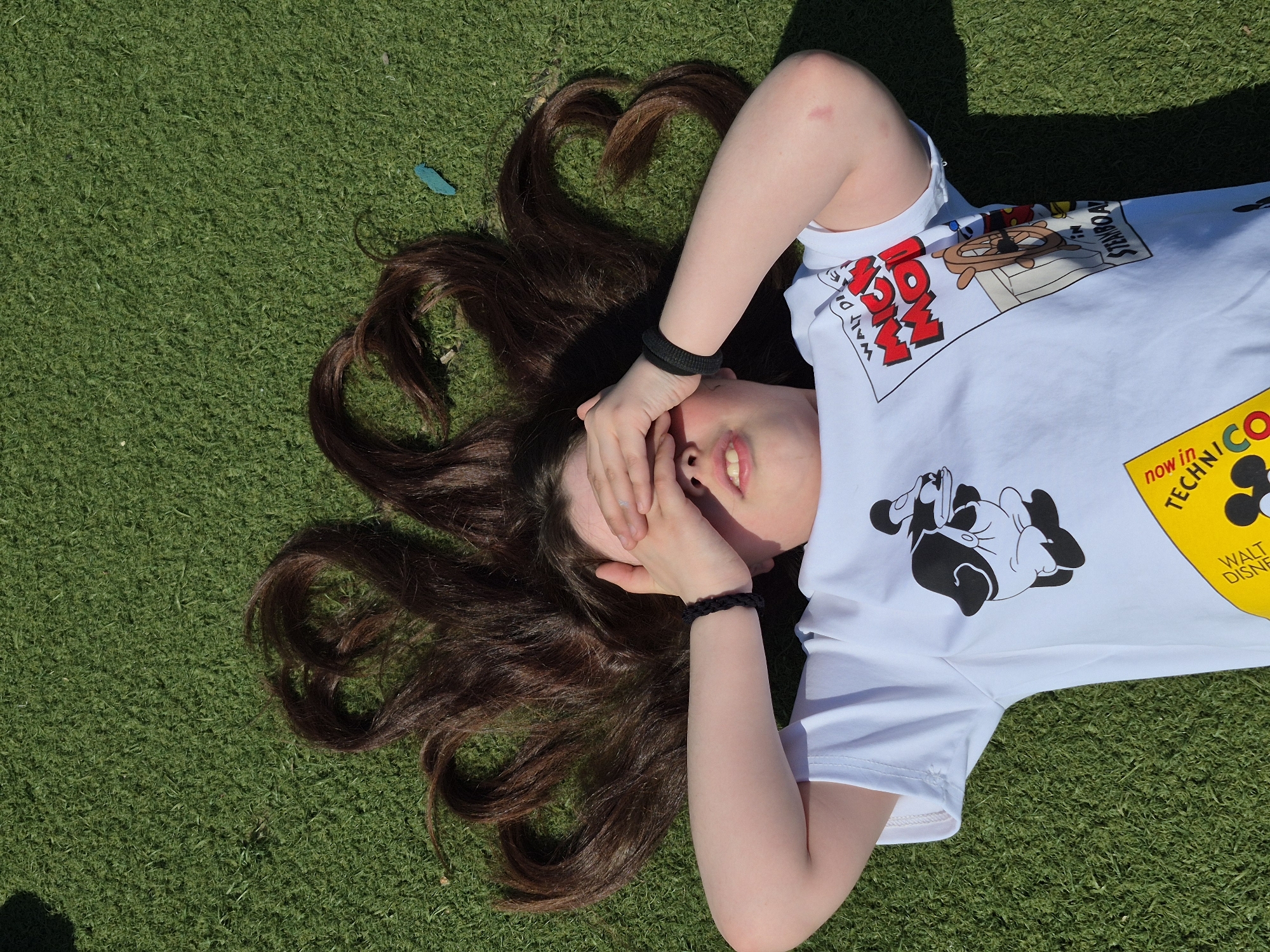 A young woman lies on a green grassy surface, possibly synthetic turf, outdoors. She is positioned with her head towards the left of the frame, obscuring her face with both hands, and her body extends diagonally towards the upper right corner. Her long brown hair is spread out around her head on the grass, creating a radial pattern. She wears a white t-shirt with cartoon images and text printed on it, including depictions of Disney characters such as Mickey Mouse and wording like "Walt Disney". The sun is casting shadows on the right side of the frame, indicating a strong light source. A small, blue, unidentified object is visible on the grass near her hair. The location is not explicitly identifiable as the city of Chaikovsky, Russia, based on the provided visual information.