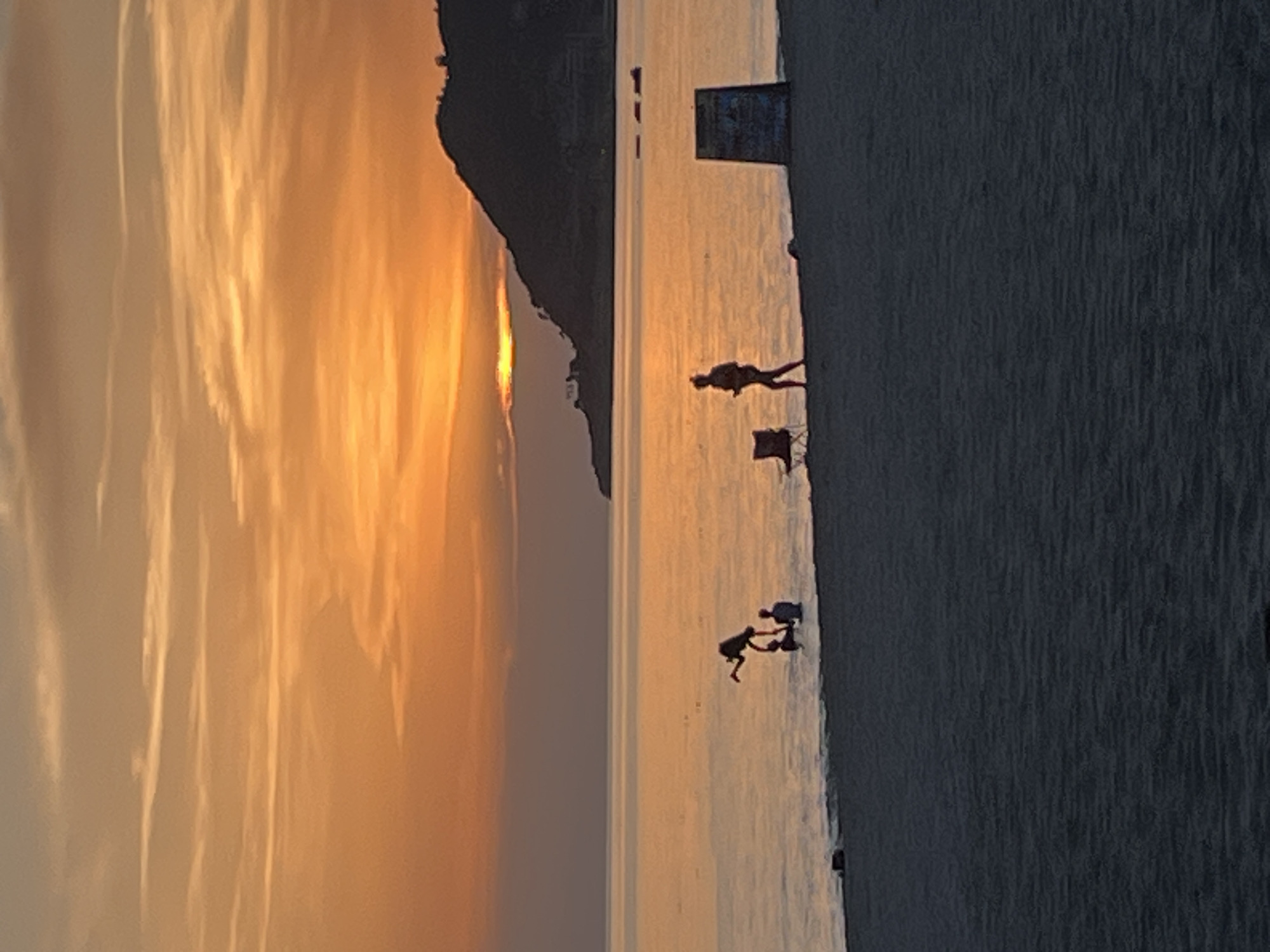 The image depicts a coastal scene in Alanya, Turkey, at sunset. The sky is streaked with orange and yellow hues, reflecting on the calm sea. Silhouetted figures are visible on the sandy beach. One individual is standing, with a chair nearby. Further down the beach, two smaller figures, possibly children, are interacting near the water's edge. In the background, a dark headland juts out into the sea. A rectangular structure stands on the beach further up. The image suggests a peaceful, relaxed atmosphere, typical of a beach scene at dusk.