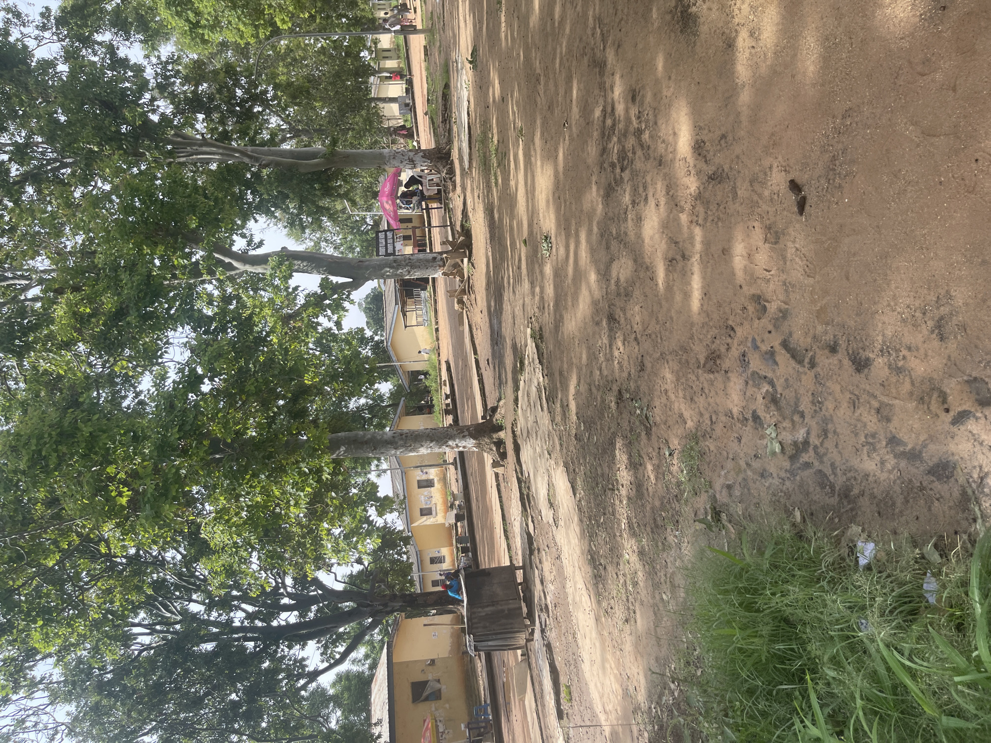 A wide shot shows an outdoor scene in Rafin Par, Nigeria. The foreground is a dusty, reddish-brown dirt path with sparse patches of green grass and some scattered leaves. In the mid-ground, a collection of low, yellow buildings with corrugated metal roofs is visible, set back from the path. Some of these buildings appear to be small shops or residences, with windows and open doorways.
Several large trees with full green canopies dominate the scene, casting dappled shadows on the ground. One tree in the right mid-ground has a white, textured trunk. In front of one of the yellow buildings, a pink umbrella provides shade over a small seating area where two individuals are seated. Nearby, a gray metal trash receptacle stands prominently in the center of the frame. A person wearing a blue shirt and a red cap is visible near the trash receptacle.
Further back, more of the yellow buildings and trees are visible, suggesting a village or a small town setting. A few other individuals are seen walking in the distance. The overall impression is one of a casual, everyday scene in a warm, outdoor environment. The sky is overcast and bright.