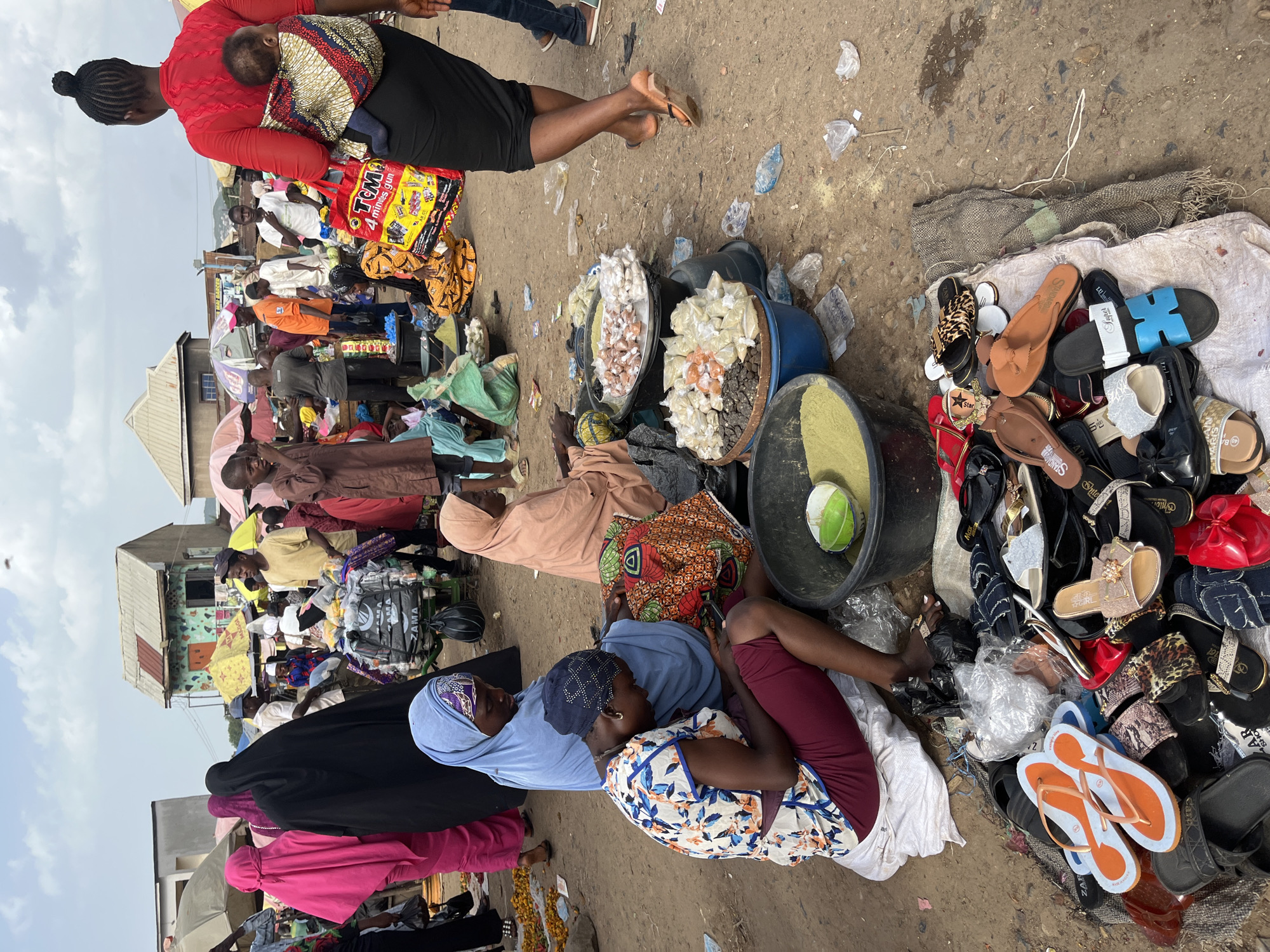A bustling outdoor market scene unfolds in Rafin Par, Nigeria. In the foreground, a collection of footwear, including flip-flops and sandals, is displayed on the ground. Further back, several individuals are engaged in commerce. A woman in a red top carries a baby on her back, holding a large shopping bag. Another woman in a black hijab is seen from behind, also with a baby on her back. A woman in a light blue hijab is sitting, looking down at her phone, while another woman in a peach-colored hijab sits beside her. A vendor is arranging food items, possibly spices or grains, in large bowls and bags. The market appears to be a vibrant hub of activity, with people dressed in a variety of traditional and contemporary clothing. The ground is dusty, indicative of an outdoor setting, and the sky is overcast. Structures resembling market stalls or small buildings can be seen in the background, with awnings providing shade. The overall impression is one of a lively and active marketplace in a Nigerian city.