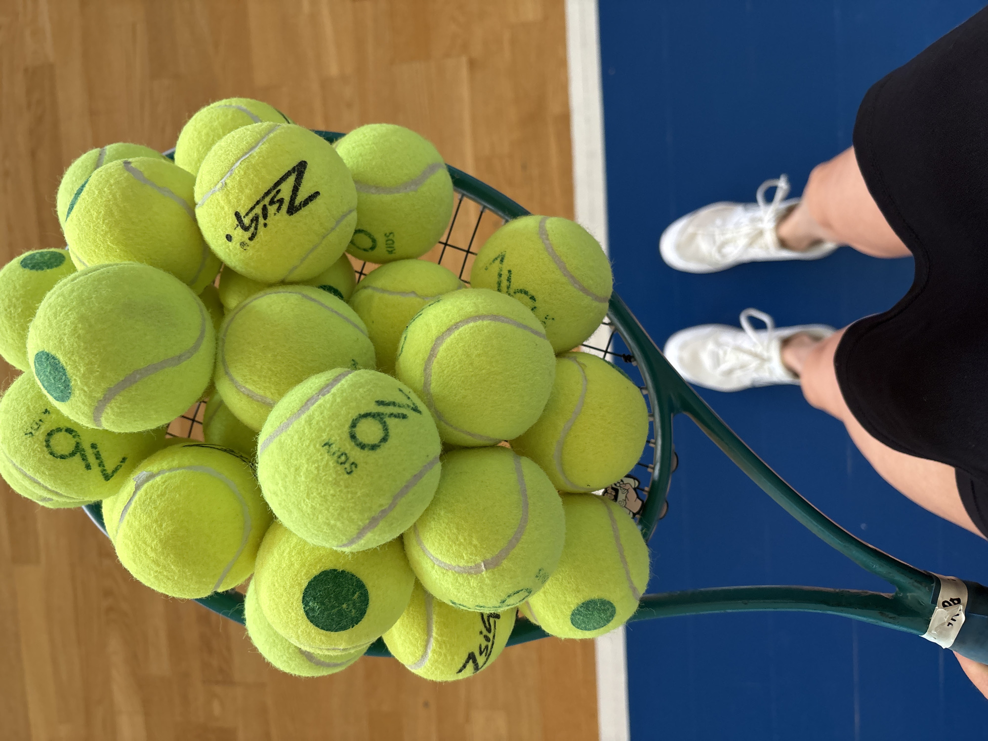 A top-down shot shows a tennis racket filled with about 20 bright yellow tennis balls labeled "KIDS" and some with markings. A person wearing white sneakers, black shorts, and a tan complexion is holding the racket. The background is divided, with one side appearing as wood flooring and the other side a blue surface marked with a white line. Based on the writing on the tennis balls being in Russian and other hints, the location of the image could be a tennis court in Chaikovsky, Russia.