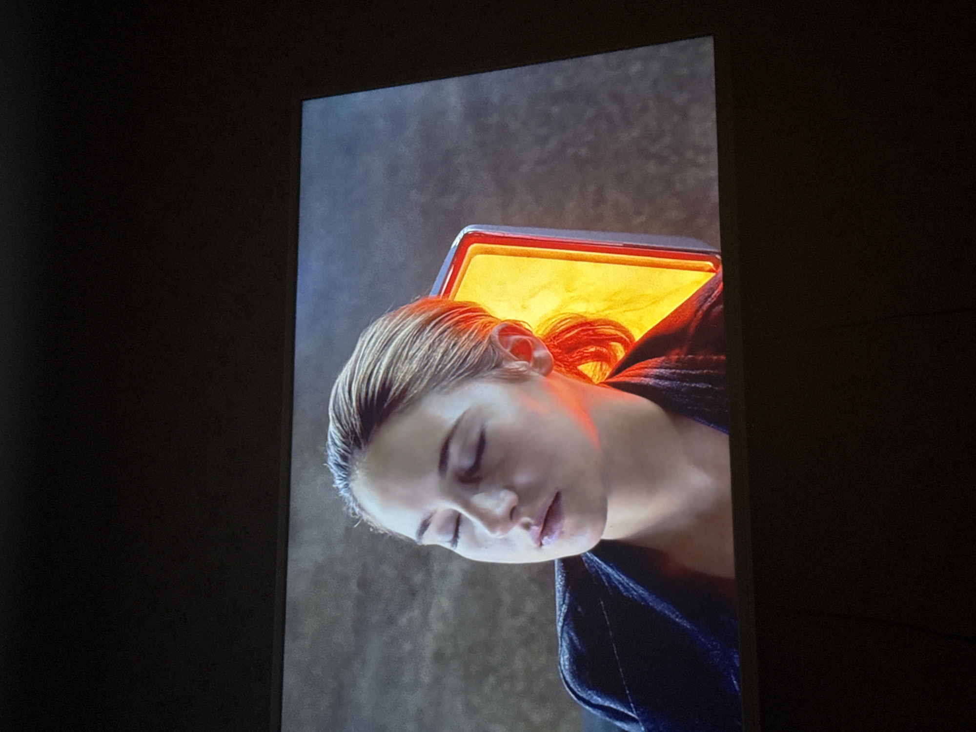 A screen displays an image of a woman, likely Shailene Woodley, undergoing what appears to be a medical or cosmetic procedure involving a light box. The woman has light hair and closed eyes. The light box emits a warm, orange glow. She is wearing a dark garment, possibly a robe. The background of the image suggests an indoor setting with a grey wall. The photograph was taken indoors. I cannot provide the location in the city of Mytishchi, Russia, as the image does not contain that information.