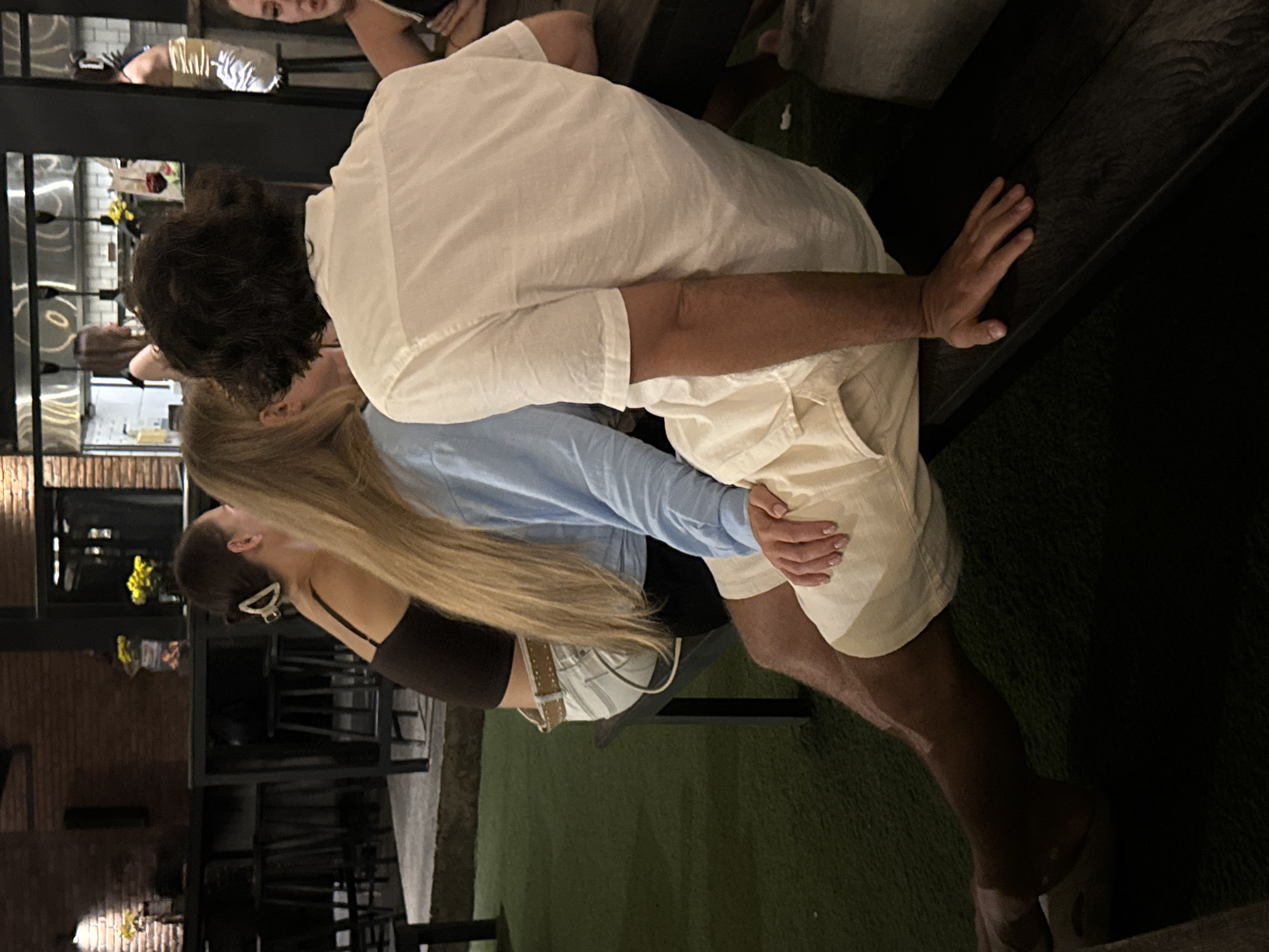 The image shows a man and a woman in an embrace. The man is wearing a white shirt and light-colored shorts. The woman has long blonde hair and is wearing a black top and blue long-sleeved shirt. They are standing outdoors, with a brick building visible in the background. The location is identified as North Kuta, Indonesia.