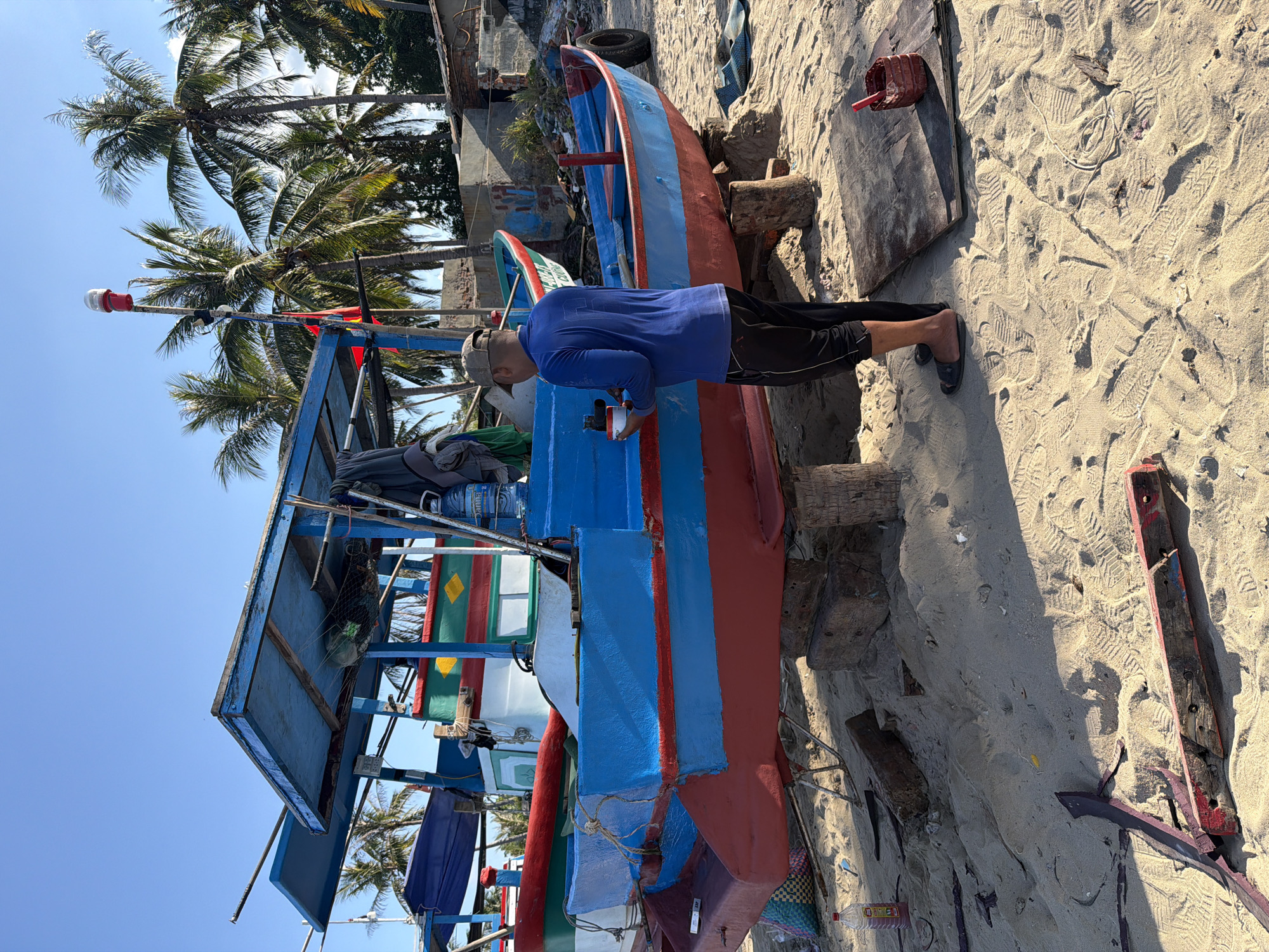 The image shows a man working on a blue and red boat on a sandy beach. The boat is propped up on wooden blocks. The man is wearing a blue shirt, dark shorts, and slippers, and has a cap on his head. He appears to be holding something in his hands and working on the side of the boat. In the background, there are palm trees and a clear blue sky. The location is in Bac Nha Trang Ward, Vietnam. There are other boats nearby. The scene also contains various objects like wooden planks, a plastic bottle, and a red container scattered on the sand.