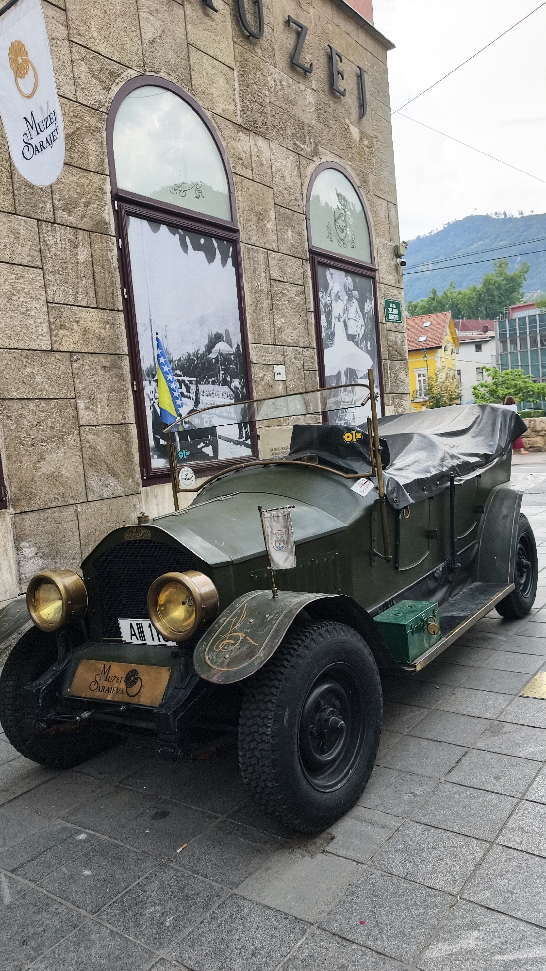 The image shows a vintage green car parked on a cobblestone street in front of a building with a stone facade. The building has arched windows displaying historical images and a sign that reads "MUZEJ SARAJEVO". A Bosnian flag is displayed on the car. The car appears to be an exhibit, possibly at a museum. The street is paved with grey square stones. In the background, there are other buildings and a hillside with trees and houses. The sky is overcast.