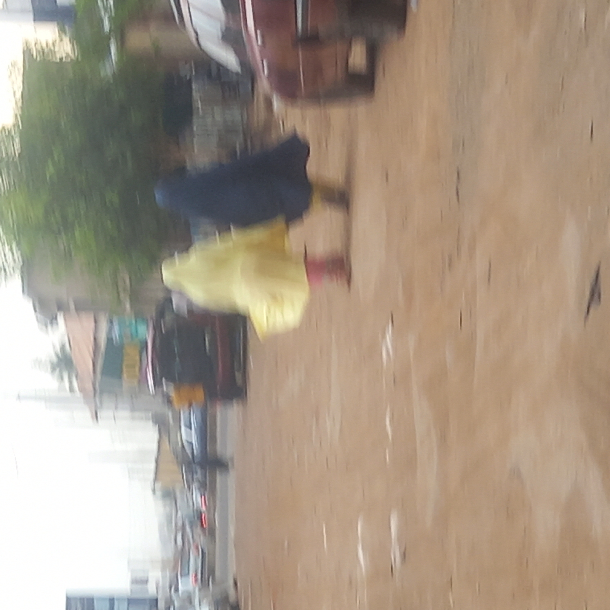 This heavily blurred image captures an outdoor scene, likely a street or unpaved road, in Dadi Riba, Nigeria. The ground is a light brown, sandy surface.
The primary subjects are two people, seen from behind, walking away from the camera. The figure closer to the foreground is wearing a bright yellow garment, possibly a loose dress or traditional abaya, and appears to have red footwear. Partially obscured behind and to the left of this person is another figure dressed in a dark blue or black garment, also possibly a traditional outer layer. Both individuals are in motion, contributing to the significant blur.
In the mid-ground and background, several vehicles are visible on the left side of the road. One prominent vehicle appears to be a dark red or maroon sedan or SUV. Green foliage, likely trees or bushes, lines the background to the left of the road, behind the vehicles. Further in the distance, indistinct buildings can be faintly seen.
The scene takes place during daylight hours, but the exact time of day or weather conditions cannot be determined precisely due to the image's blurriness and lack of clear light sources or shadows. No specific location cues beyond general street elements or discernible text are visible. The expressions or emotions of the individuals are not apparent.