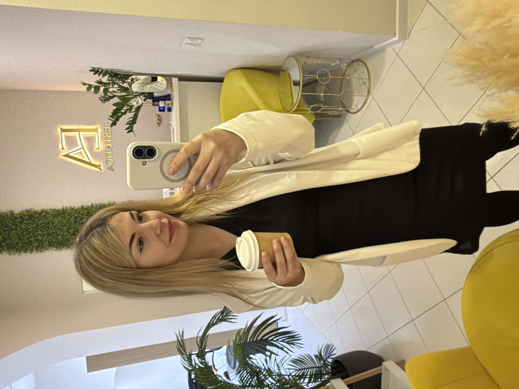 The image shows a woman holding a coffee cup and a phone in a room in Budva, Montenegro.
The woman is blond with fair skin and is wearing a white jacket over a black outfit. She holds a paper coffee cup in her left hand and a white smartphone in her right, apparently taking a selfie in a mirror.
The room is decorated in neutral tones with yellow accents. A wall is covered in greenery. A sign reading "Estetica" is illuminated in a gold color. Behind the woman are a yellow chair, a gold wire side table, and other decorative items. There are houseplants in the lower corners of the frame, and tile flooring.