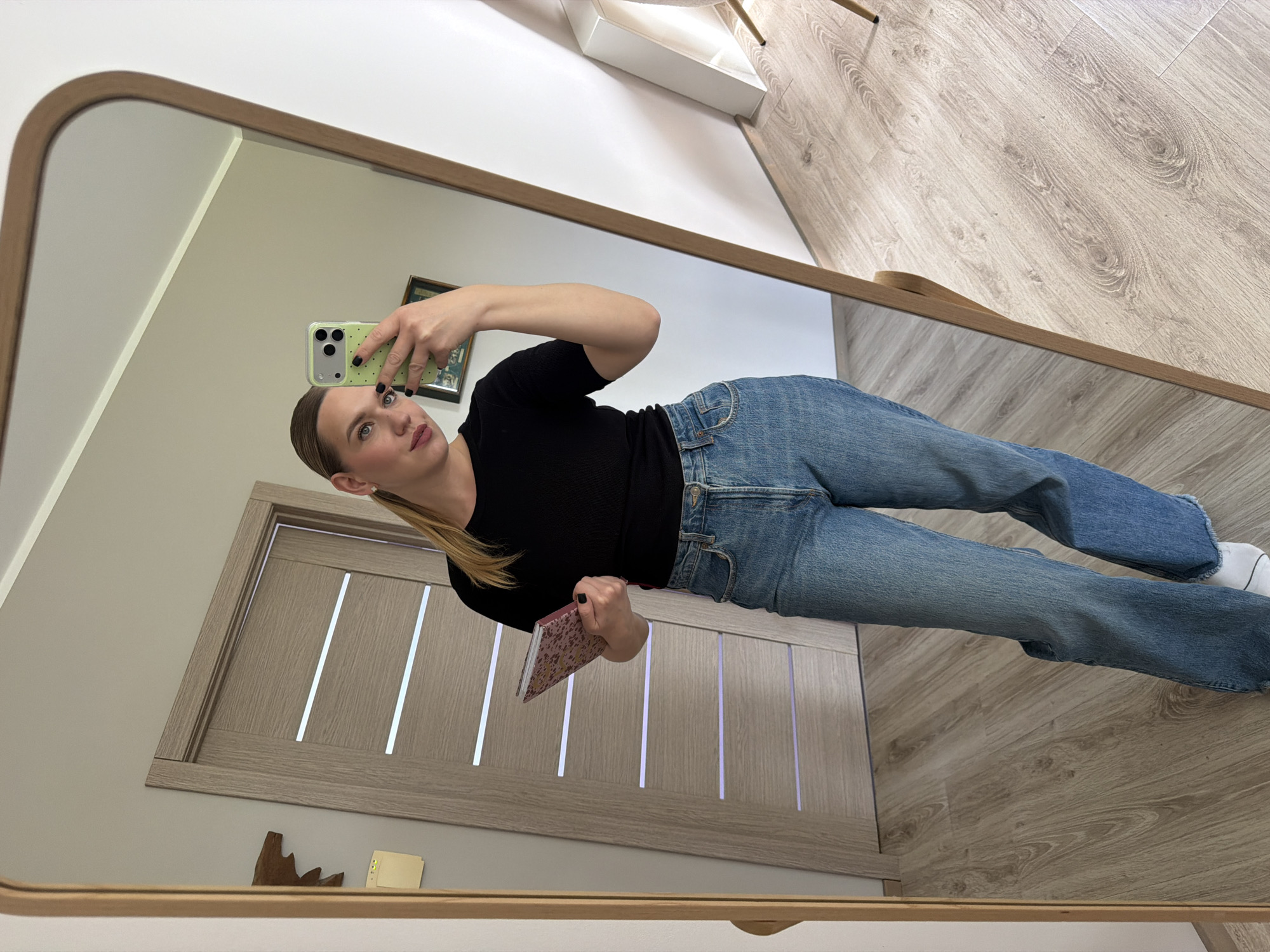 A woman is taking a selfie in a mirror. She is wearing blue jeans and a black shirt. She is holding a phone in her right hand and a book in her left hand. The reflection shows a door and a wooden floor. The location is likely Dreiliņi, Latvia, given the prompt instructions.