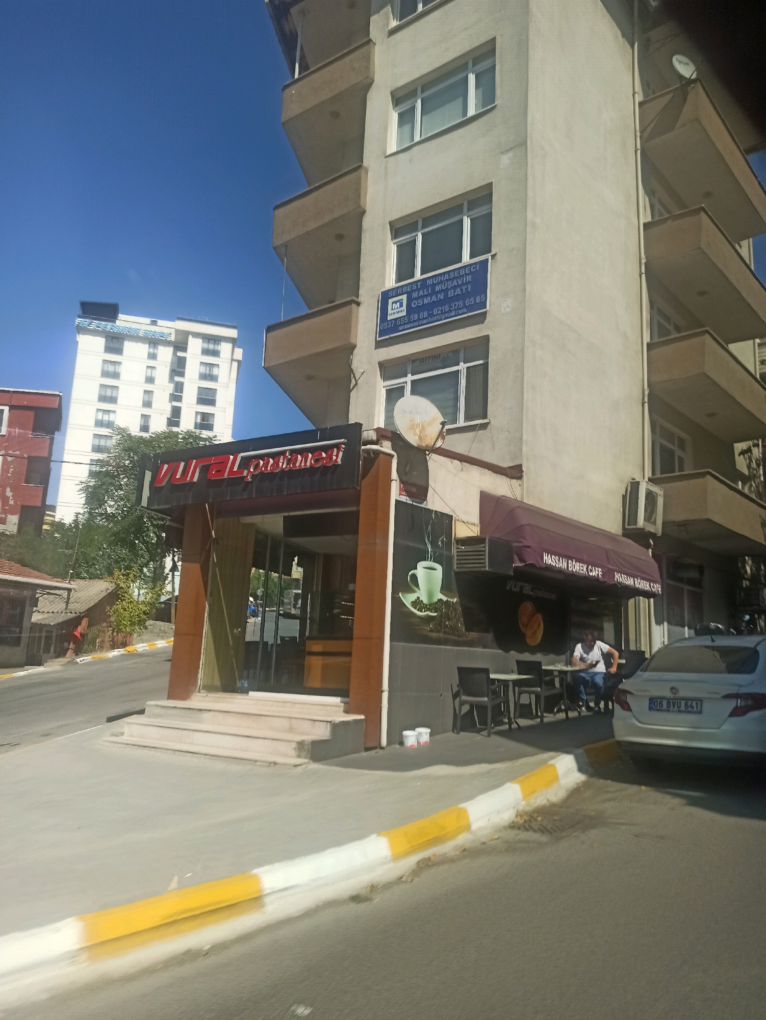 The image captures a street view in Pendik, Turkey, under a clear blue sky. In the foreground, a yellow and white striped curb separates the sidewalk from the road. A light-colored car with license plate "06 BYU 641" is parked on the right side of the road, facing away from the camera.
The mid-ground features a building with a burgundy awning displaying "HASSAN BÖREK CAFE" in white lettering. Beneath the awning, tables and chairs are set up outside the cafe. A person is seated at one of the tables, looking down at their phone. To the left of the cafe, there's a bakery with a large sign that reads "VURAL Pastanesi" in red lettering. The entrance of the bakery is a glass door. Adjacent to the bakery, a large graphic of a green cup and saucer is visible on the wall.
In the background, a tall, white residential building stands prominently. To the left of this building, another shorter, weathered red building is partially visible. The overall environment suggests a commercial and residential area within the city.
