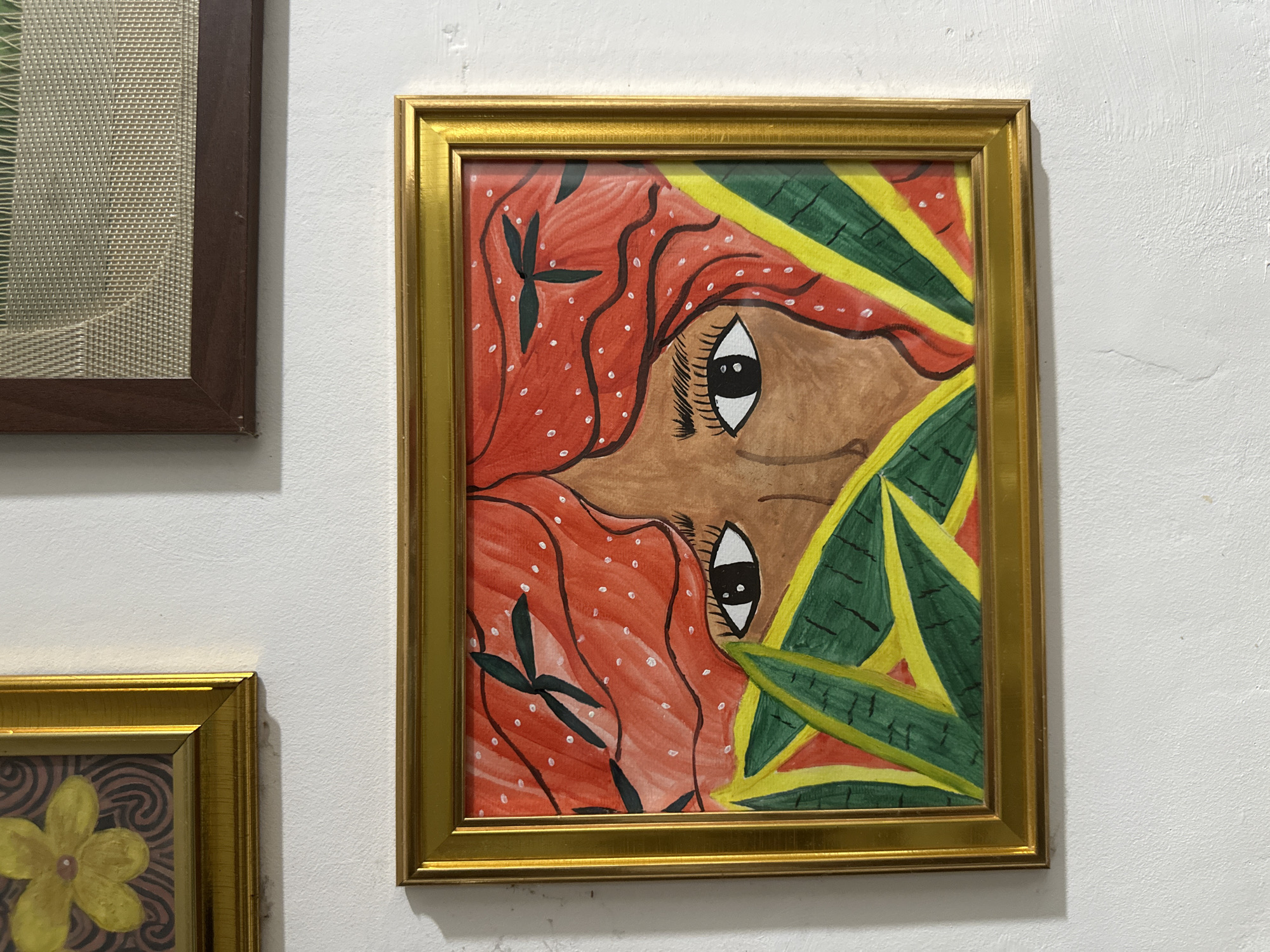 A collection of framed artworks is displayed on a textured white wall in Davao City, Philippines. The central and most prominent piece is a colorful, stylized painting presented in a gold-colored frame. The painting features a close-up of a person's face, depicted in profile and rotated 90 degrees clockwise from its intended vertical orientation. The face has brown skin, with large white eyes and black pupils, detailed with eyelashes. The hair is a vibrant reddish-orange, rendered with wavy lines and small white dots, interspersed with dark green, stylized leaf shapes. On the right side of the face, bold green leaves with bright yellow outlines and veins create a tropical or jungle-like motif, contrasting with red elements.
To the top left, a portion of another framed artwork is visible, set within a dark brown wooden frame. This artwork appears to feature a light-colored, possibly woven or textured material with a subtle pattern. Below the central painting, on the bottom left, part of a third artwork is seen in a gold frame. This piece depicts a yellow flower with a brown center, set against a dark brown background with distinct swirling patterns. The white wall itself has a slightly uneven and textured finish. No specific activity is taking place, and there is no visible text in the image.