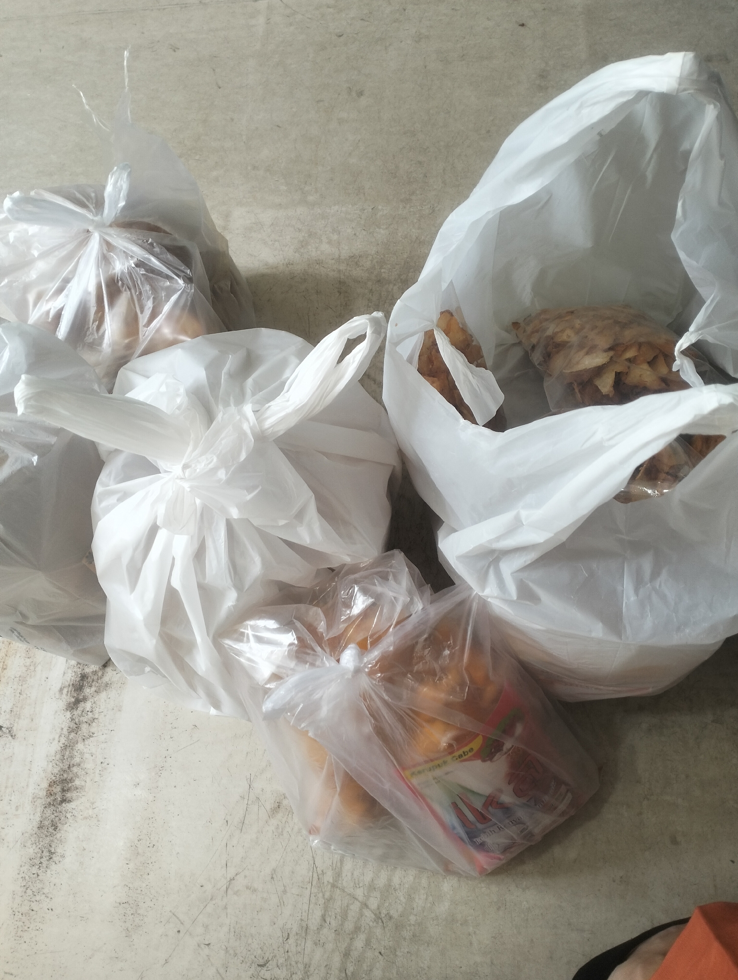 The image shows several white plastic bags filled with food items, placed on a textured, light-colored floor that appears to be concrete or a similar material. Some of the bags are tied shut, while others are open, revealing their contents. Inside the bags, one can discern what appear to be snacks or food items, some golden brown and others lighter in color, possibly including what look like chips or crackers and possibly some fried items. The lighting suggests an indoor or covered outdoor setting. The location is identified as Air Jamban, Indonesia.