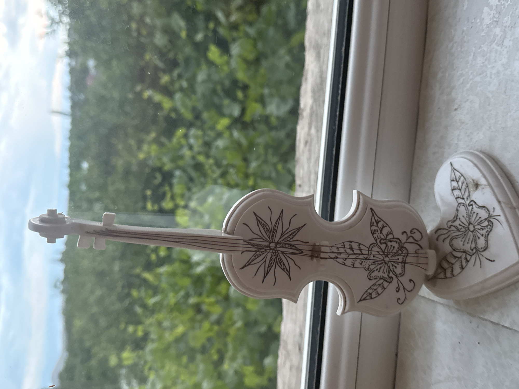 A light-colored, decorative object shaped like a violin, adorned with intricate etched floral and leaf patterns in dark lines, rests on a window sill. The violin features a star-like flower design on its upper bout and another detailed floral motif with leaves and swirls on its lower body, complete with simulated strings, a bridge, and tuning pegs. Adjacent to it, partially visible, is a matching heart-shaped ornament, also decorated with a flower and leaf pattern.
These ornamental pieces are placed on a textured, light-colored window sill, next to a white window frame with a dark inner seal. Through the windowpane, a lush outdoor scene unfolds, dominated by dense green foliage, likely trees or bushes. Above the greenery, a light blue sky with scattered white clouds is visible, suggesting daytime with overcast or partly cloudy weather. No human activity or visible text is present in the image.