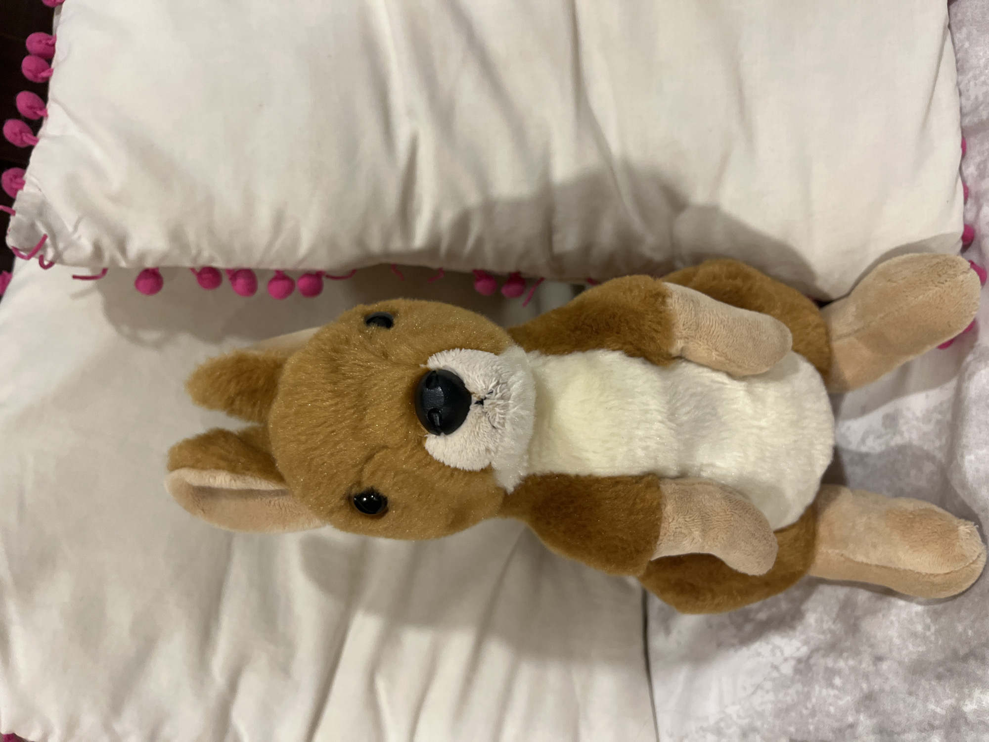 A brown and white stuffed kangaroo toy lies on its back on a white pillow with pink pom-poms. The kangaroo is positioned in the center of the frame. The background consists of the pillow and what appears to be a bedspread or a blanket with a textured pattern.
There is no location in the city of Barrio de Nuevo León, Mexico, or any people, actions or interactions present in the image.