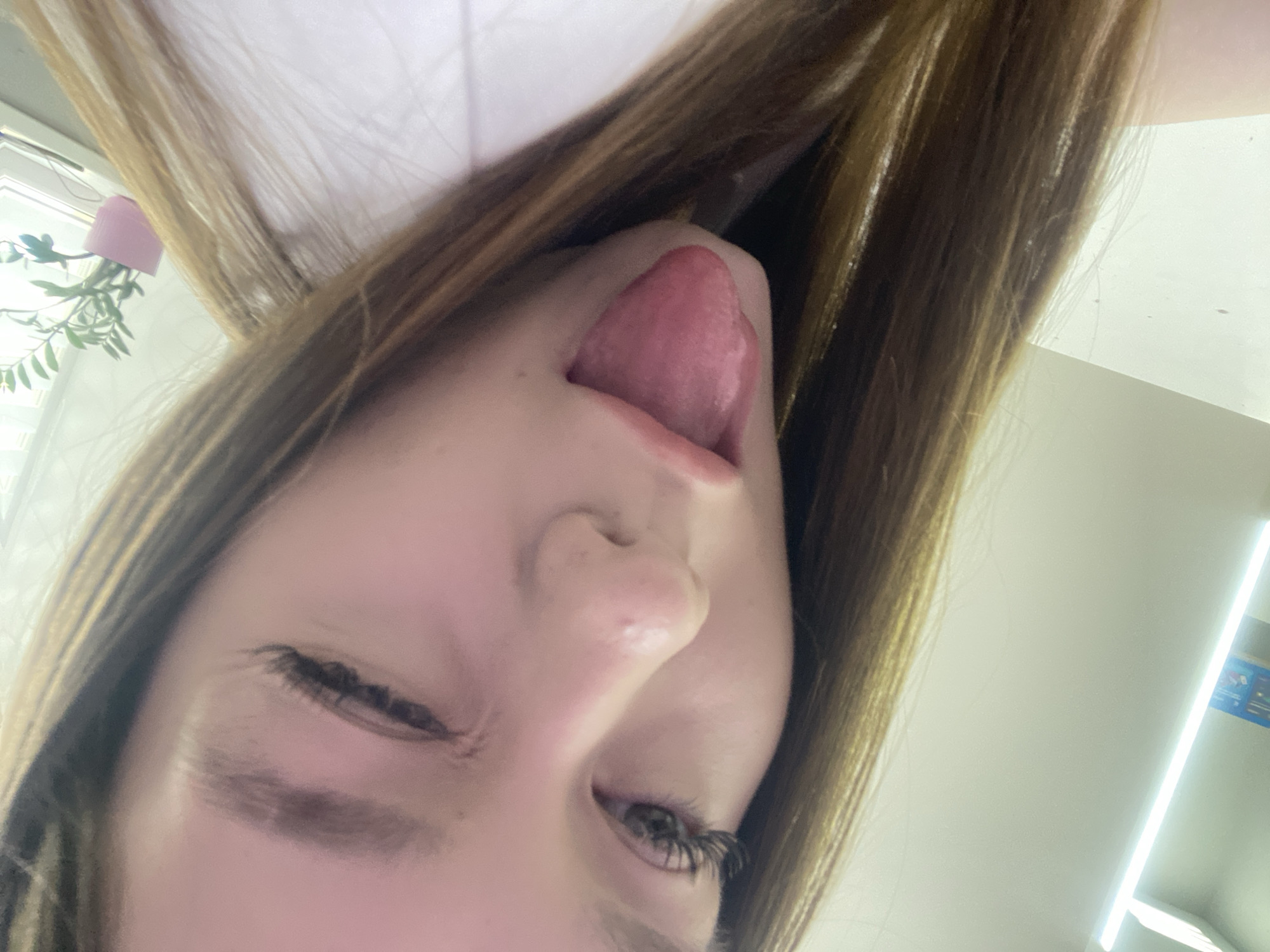 An upside-down close-up shot shows a Caucasian female with light brown hair sticking her tongue out. Her right eye is partially closed, and she is looking downward. Artificial eyelashes appear to be present. The setting is indoors, and light from a fluorescent light fixture illuminates the scene. The location is indiscernible from the image. There is no information available to determine if it is located in the city of Chaikovsky, Russia.