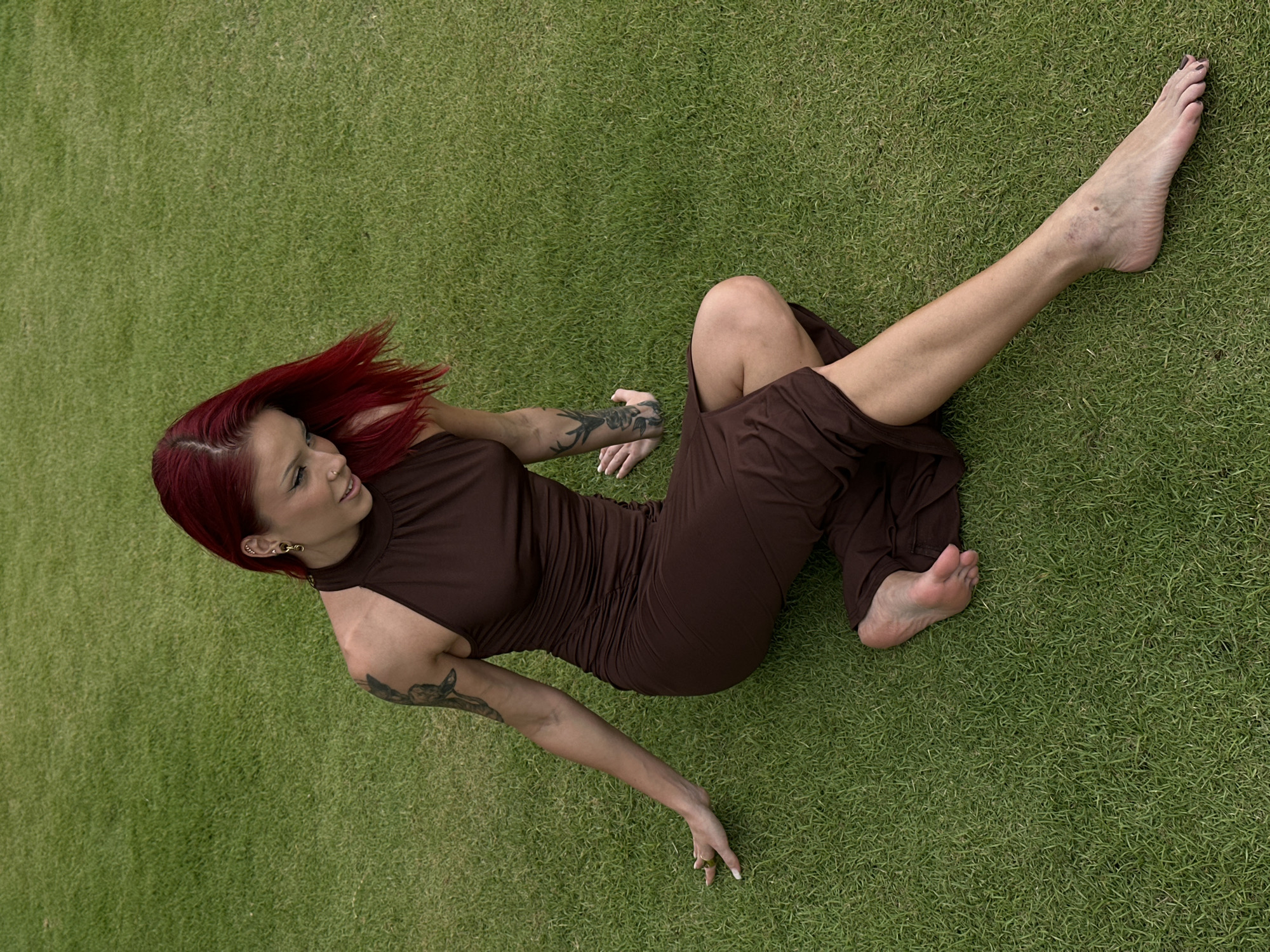 A red-haired woman in a brown dress lies on a green lawn. She is barefoot and has tattoos on her arms. She appears to be posing for a photograph. The location is likely Nusa Dua, Indonesia, based on the tropical appearance of the lawn.