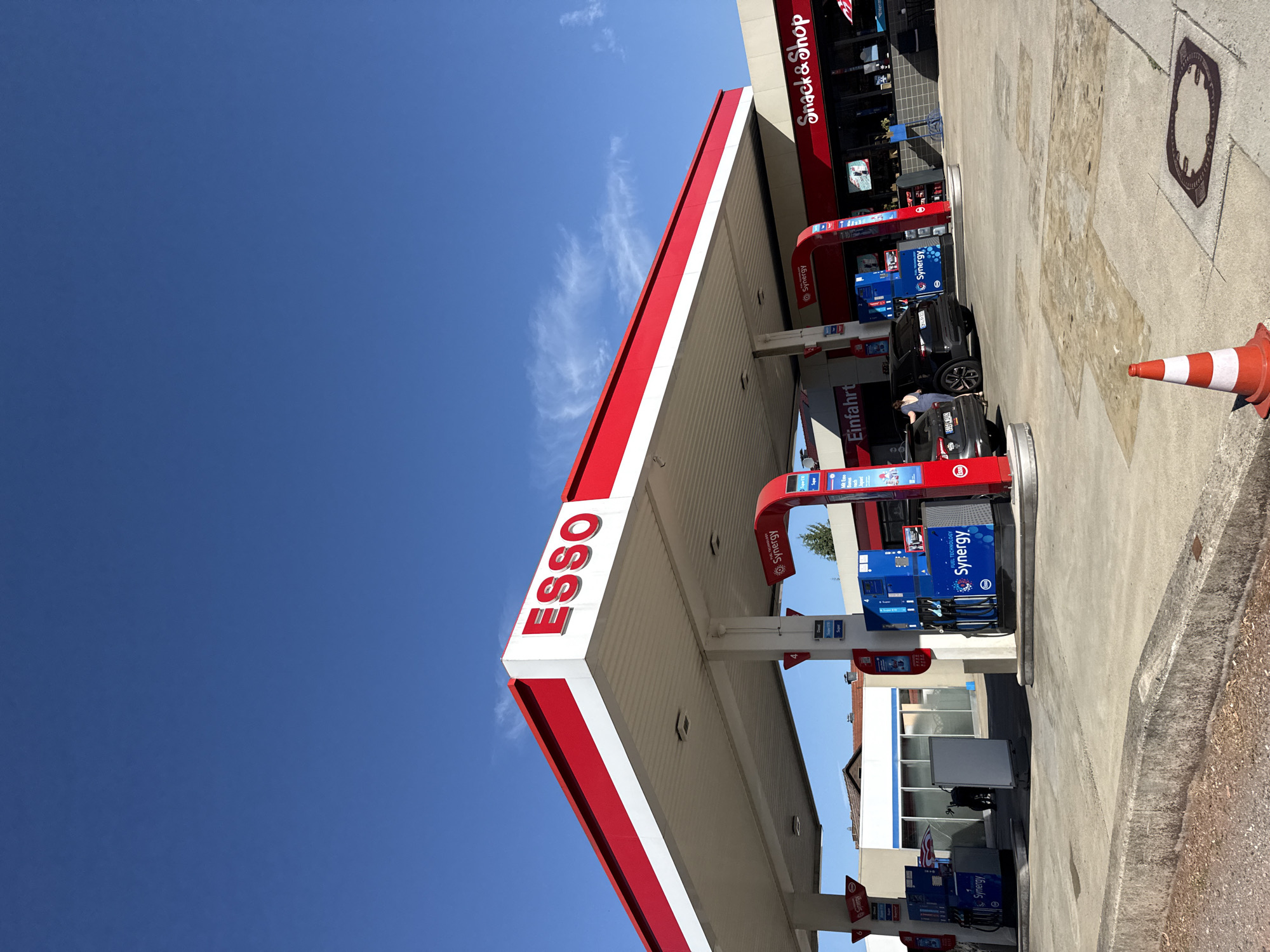 The image shows an Esso gas station with a Snack & Shop attached, located on a sunny day in Bad Sachsa, Germany. The gas station has a red and white canopy covering multiple fueling stations. The Esso logo is prominently displayed on the canopy. Several cars are parked at the fueling stations. A Snack & Shop building with a red awning is visible to the right of the gas station. In the foreground, a traffic cone is placed on the concrete ground. The sky is clear and blue with some wispy clouds. The ground appears to be concrete and asphalt. The scene is viewed from a slightly low angle, looking up towards the gas station and shop.